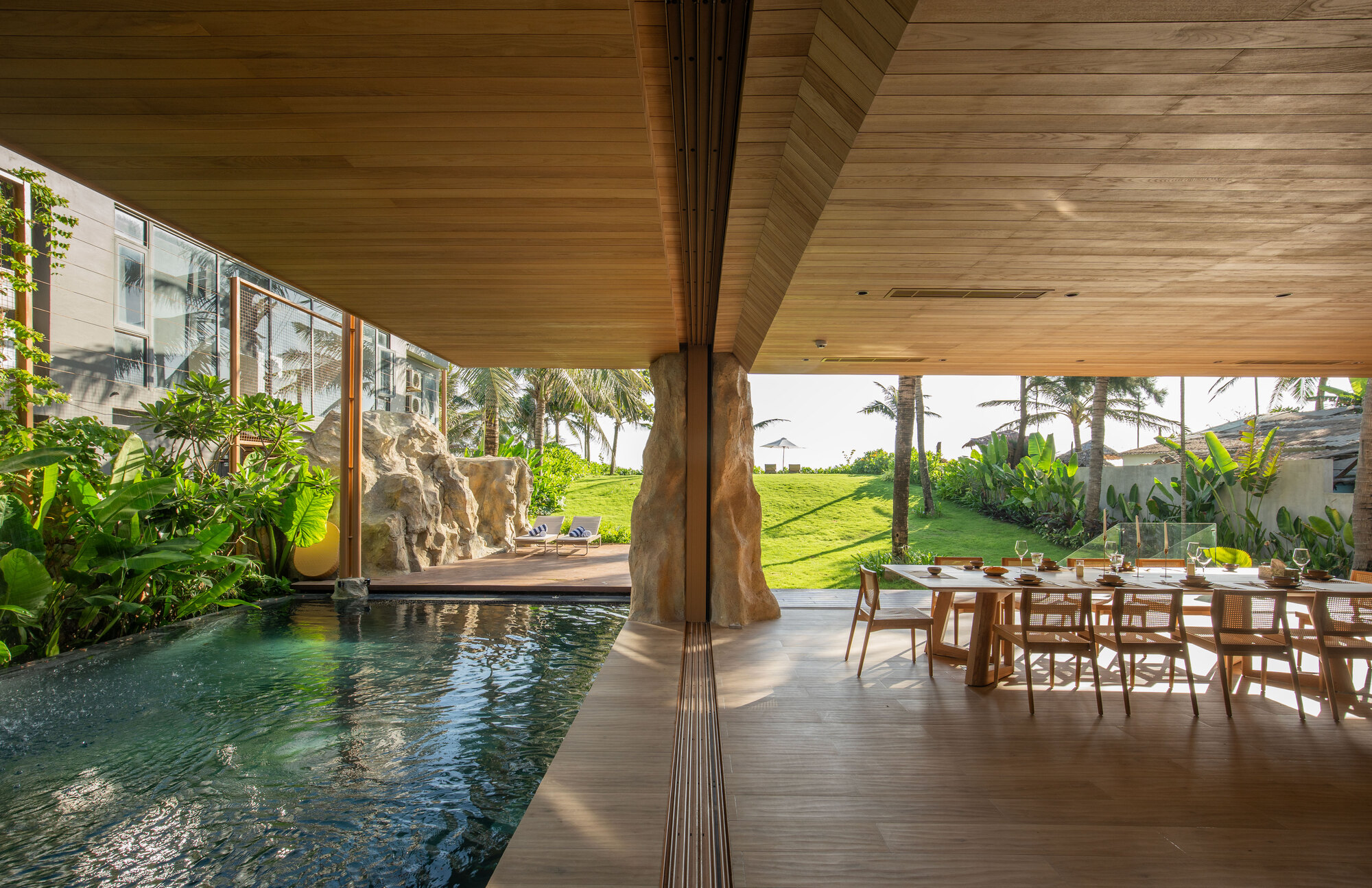 Indoor pool edge with raw stone column supporting timber ceiling beams beside dining area with tropical plants