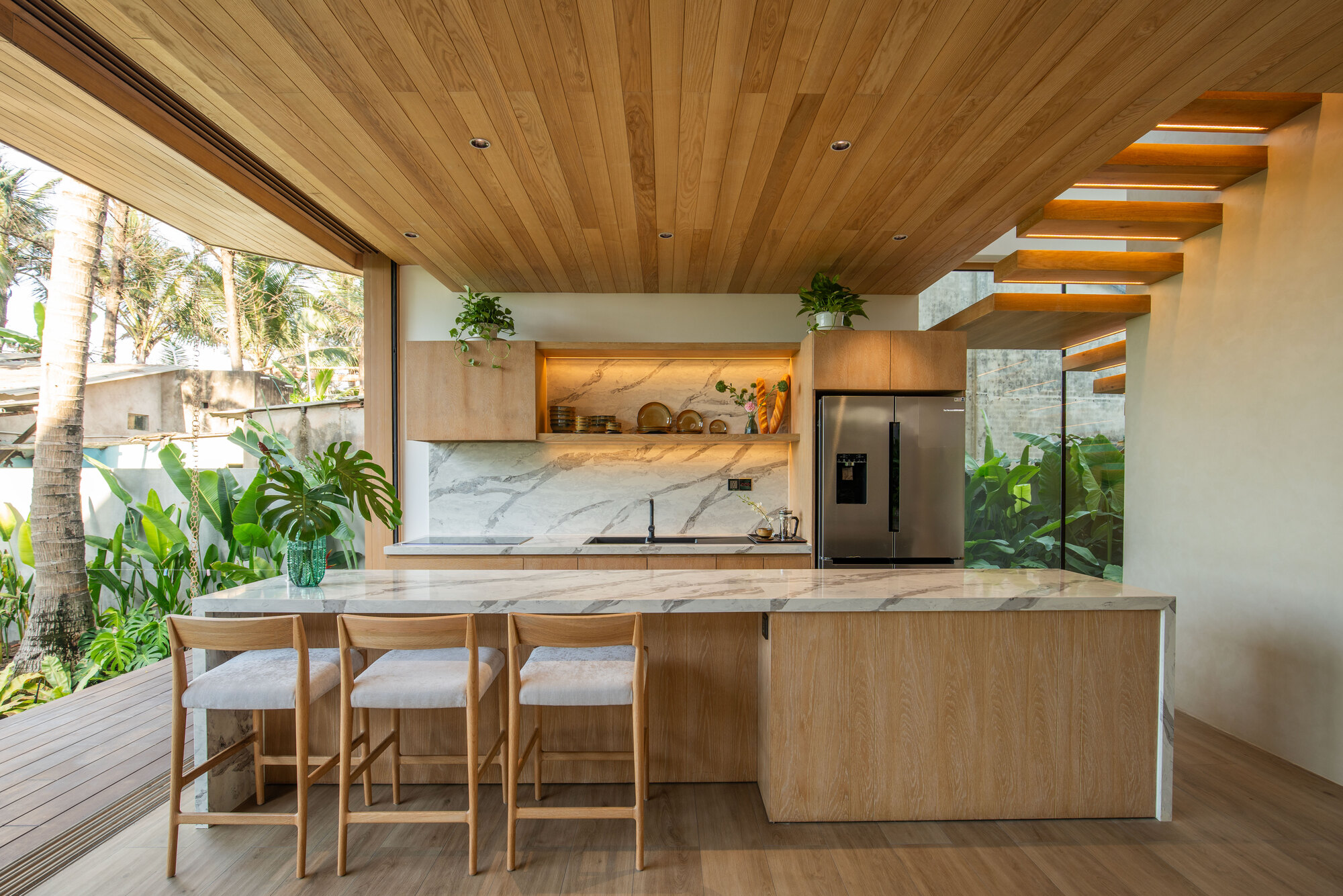 Kitchen island with marble waterfall countertop and timber cabinetry under cantilever timber staircase with plantings outside