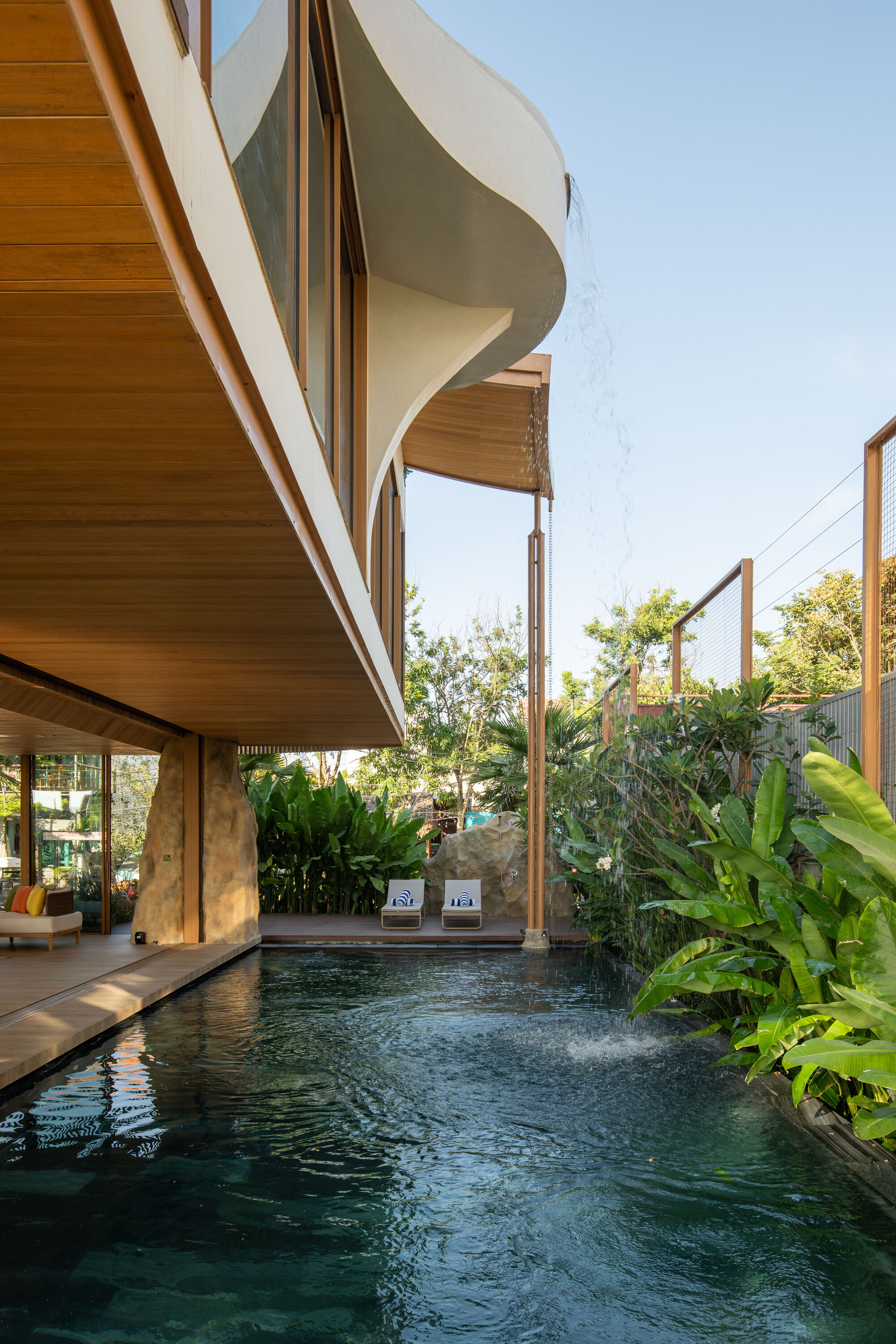 Curved upper balcony with timber soffit floating above pool terrace surrounded by tropical foliage in daylight