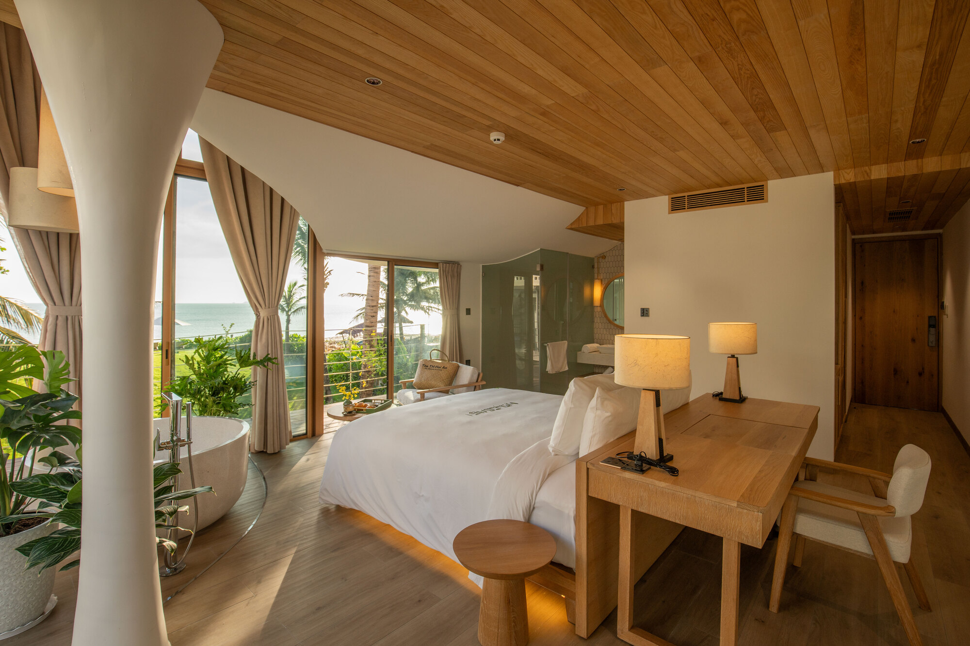 Bedroom with freestanding bathtub and timber-framed glazing opening to balcony overlooking ocean horizon at dusk