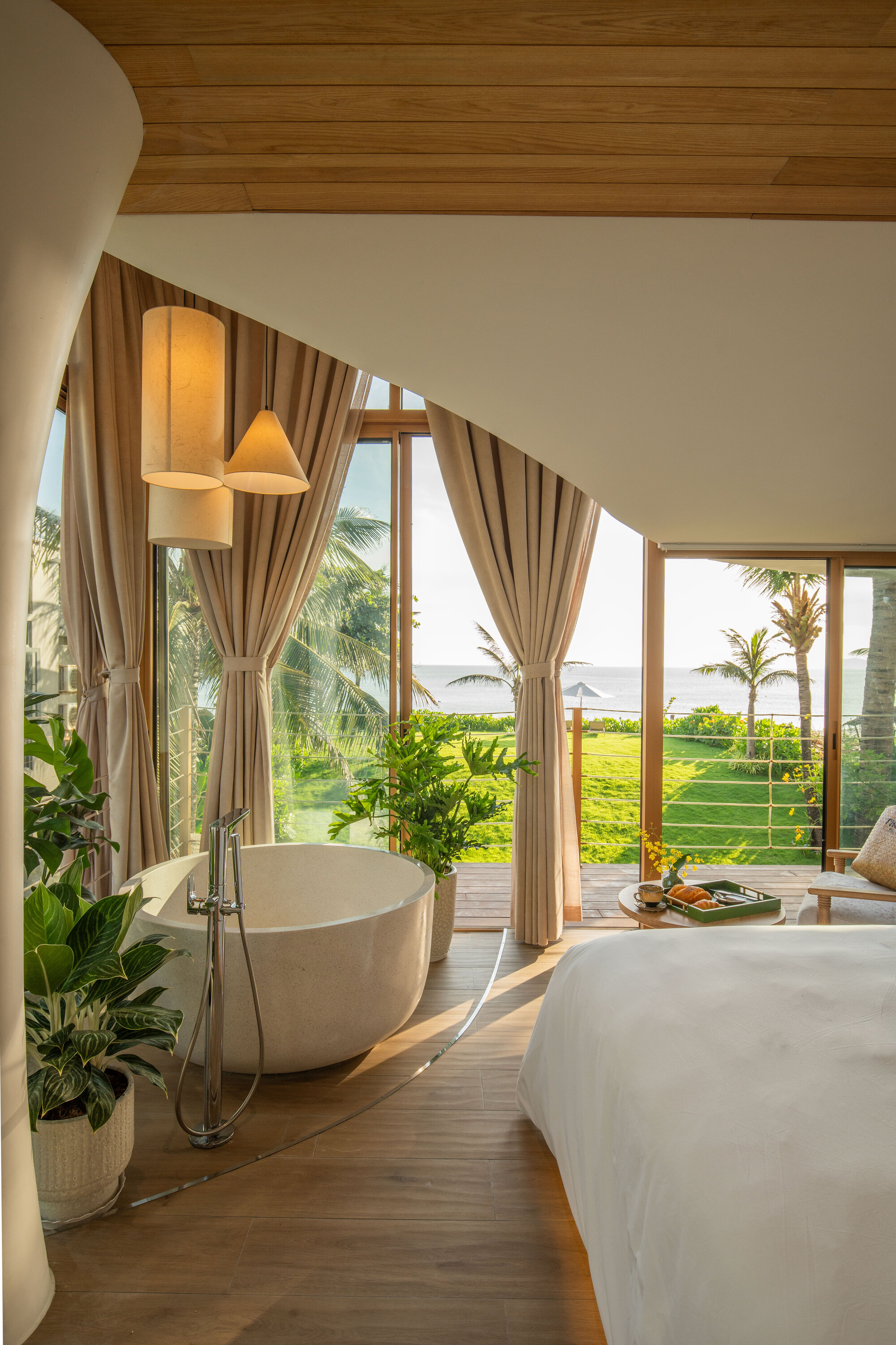 Bedroom with freestanding tub and floor-to-ceiling curtains framing ocean views with palm trees in daylight