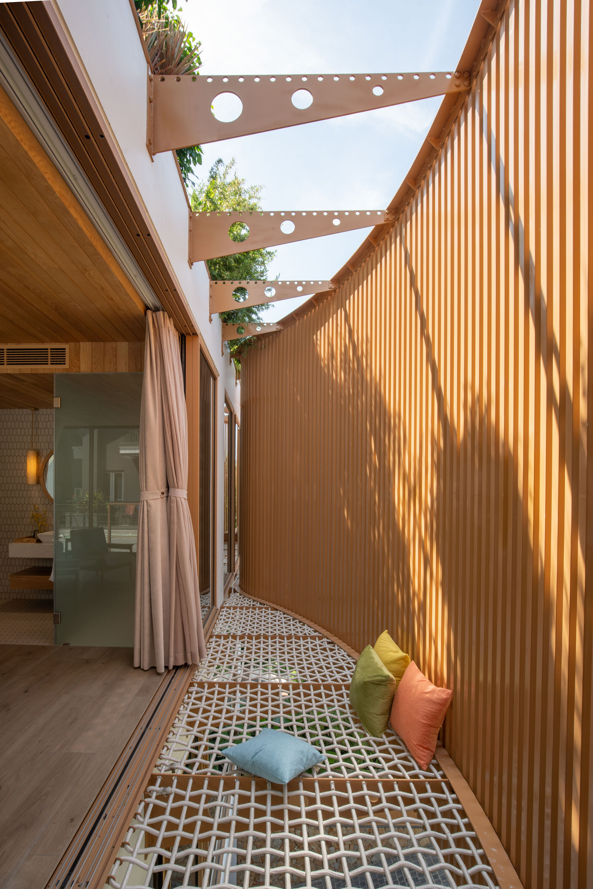 Narrow courtyard with vertical timber cladding and perforated steel beams overhead planted with greenery