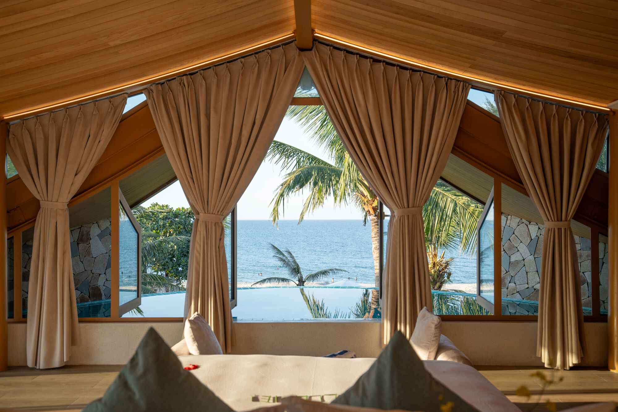 Pitched timber ceiling with clerestory windows and draped curtains overlooking infinity pool and ocean beyond