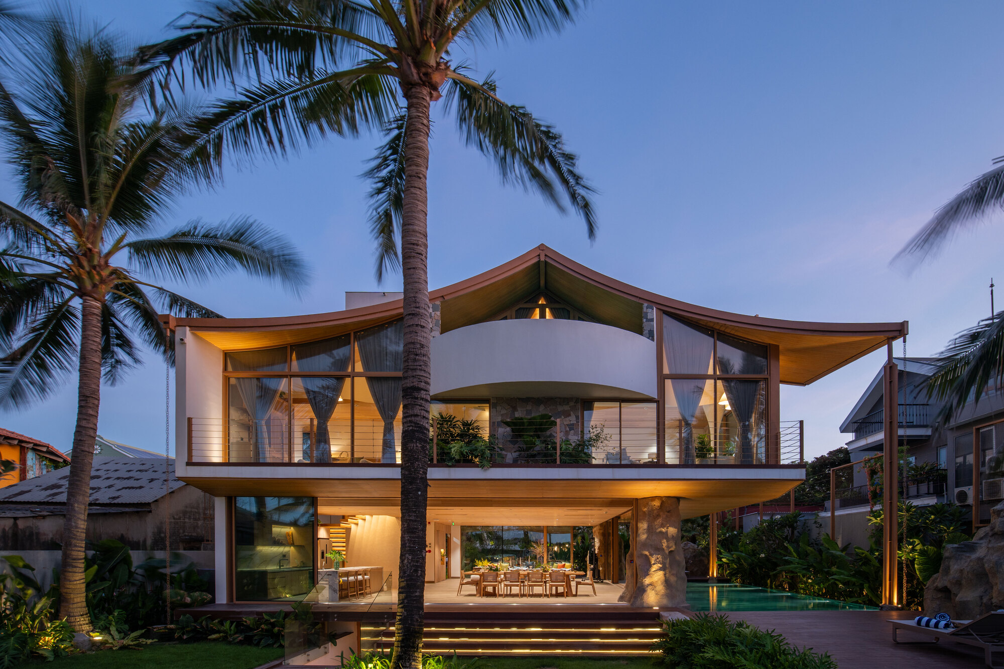 Two-story facade with curved balcony and overhanging timber eaves framed by palm trees at dusk