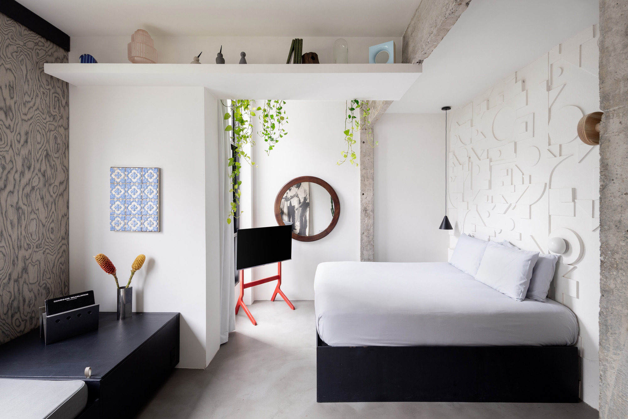 Bedroom with white textured wall panels, exposed concrete columns, and a storage shelf suspended above the bed