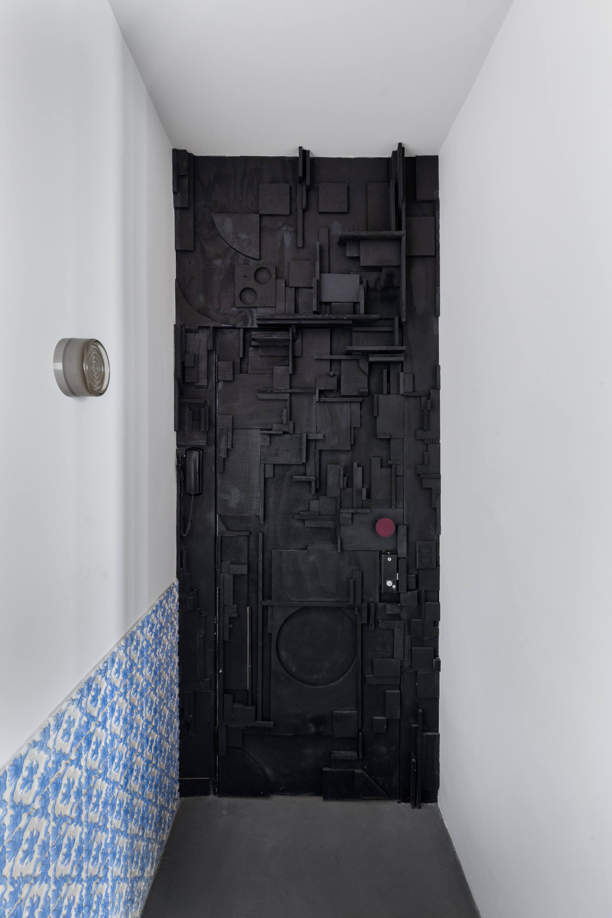 Black sculptural door composed of layered geometric forms in a narrow white corridor with blue patterned tile