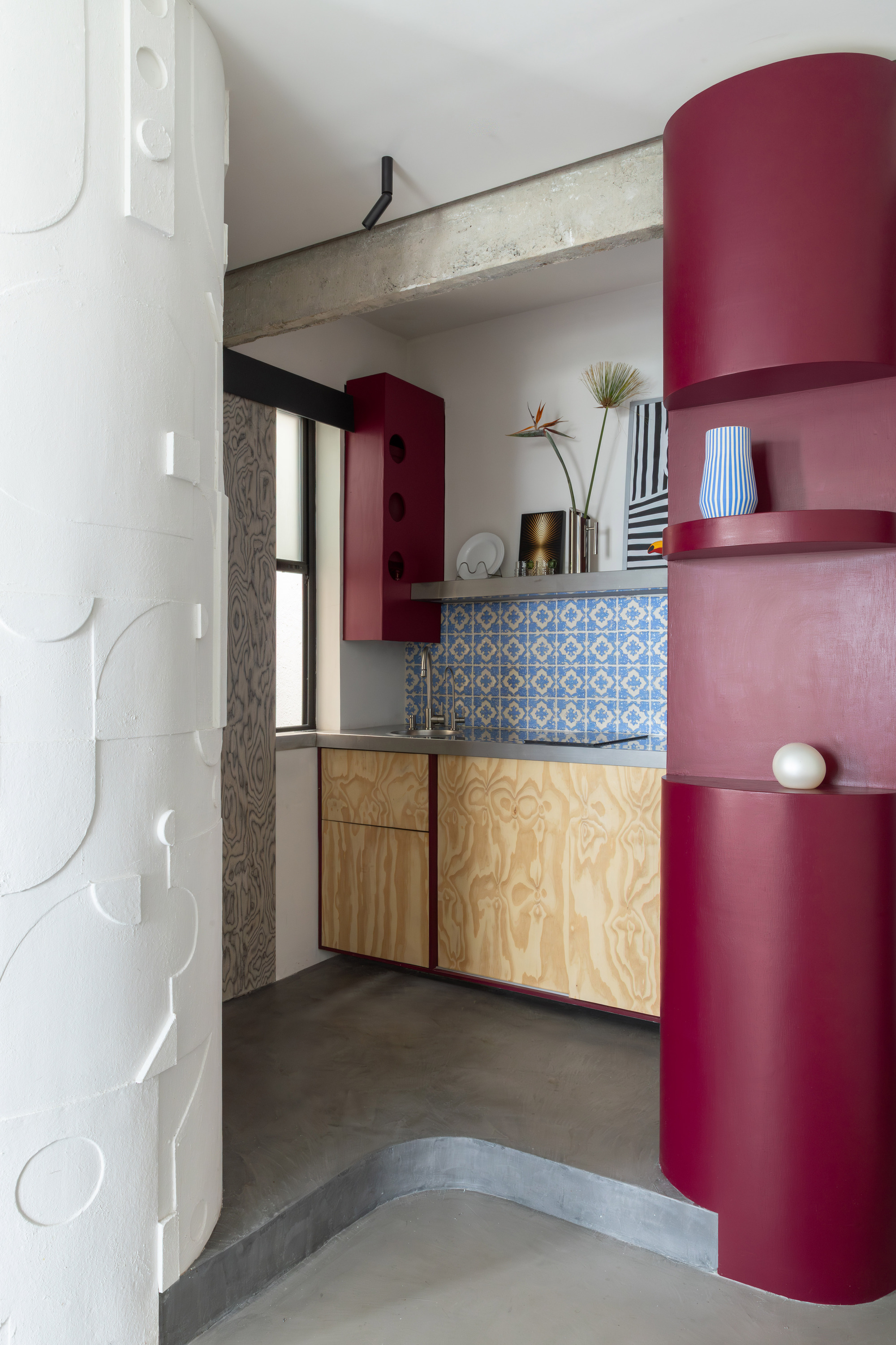 View into kitchen showing plywood base cabinets, blue tile counter surround, and exposed concrete beam overhead