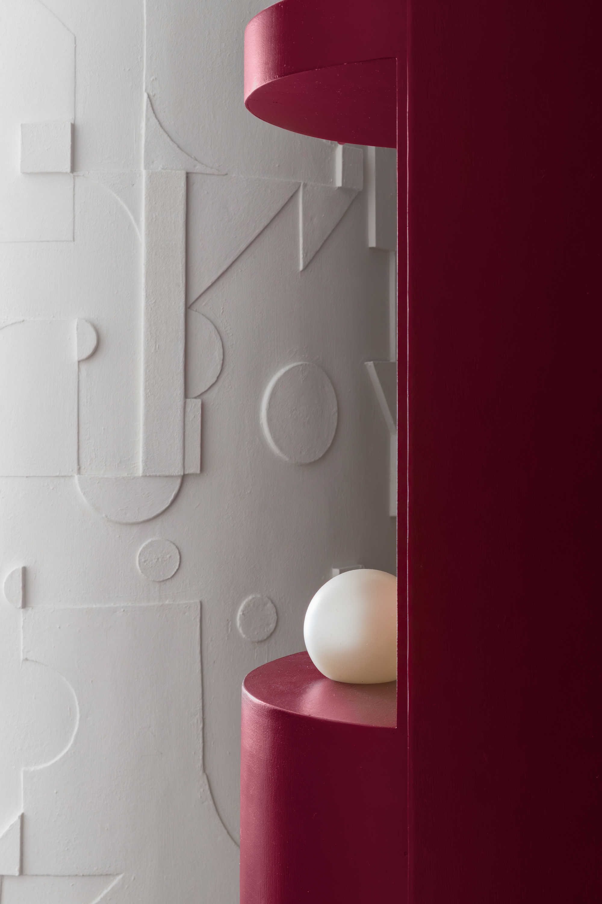 Detail of white relief wall panel with geometric forms beside a burgundy shelf holding a spherical lamp