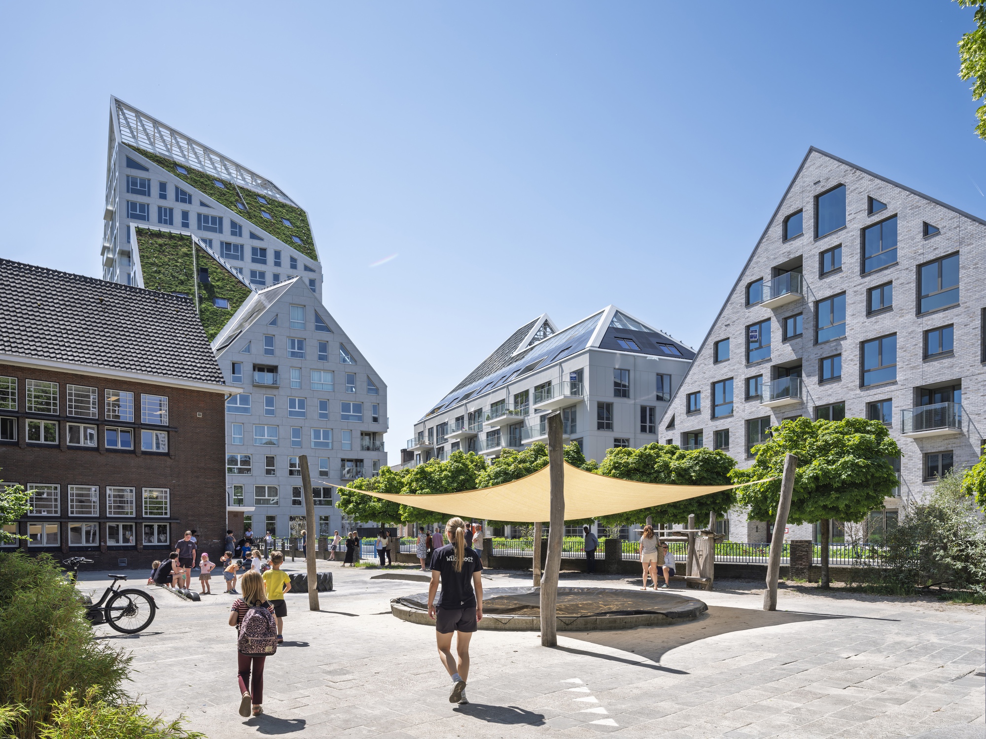 Public plaza with tensile shade canopy and residents walking among the gabled residential towers on a sunny day