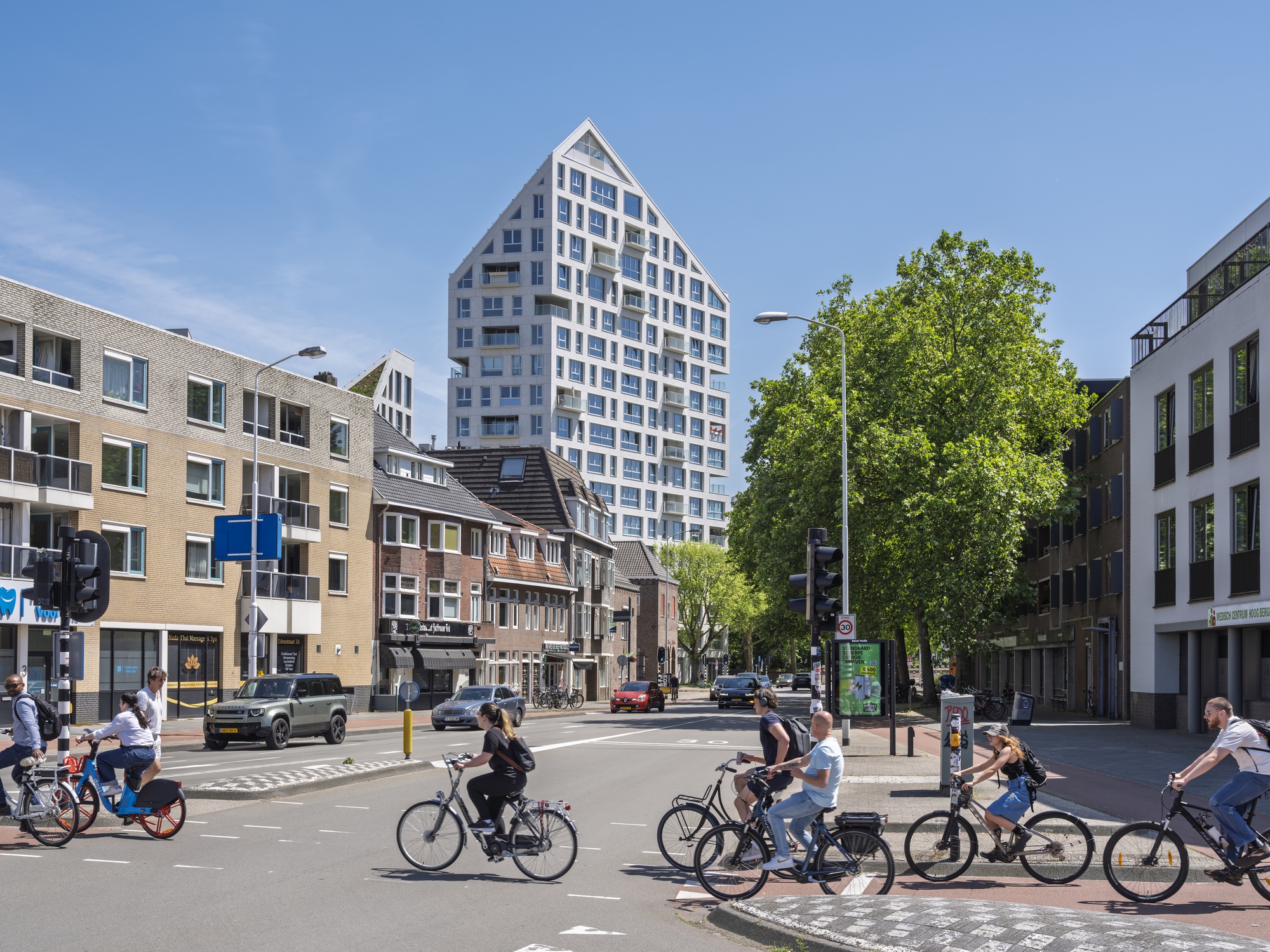 Distant view of the gabled tower from a tree-lined street intersection with cyclists crossing in the foreground