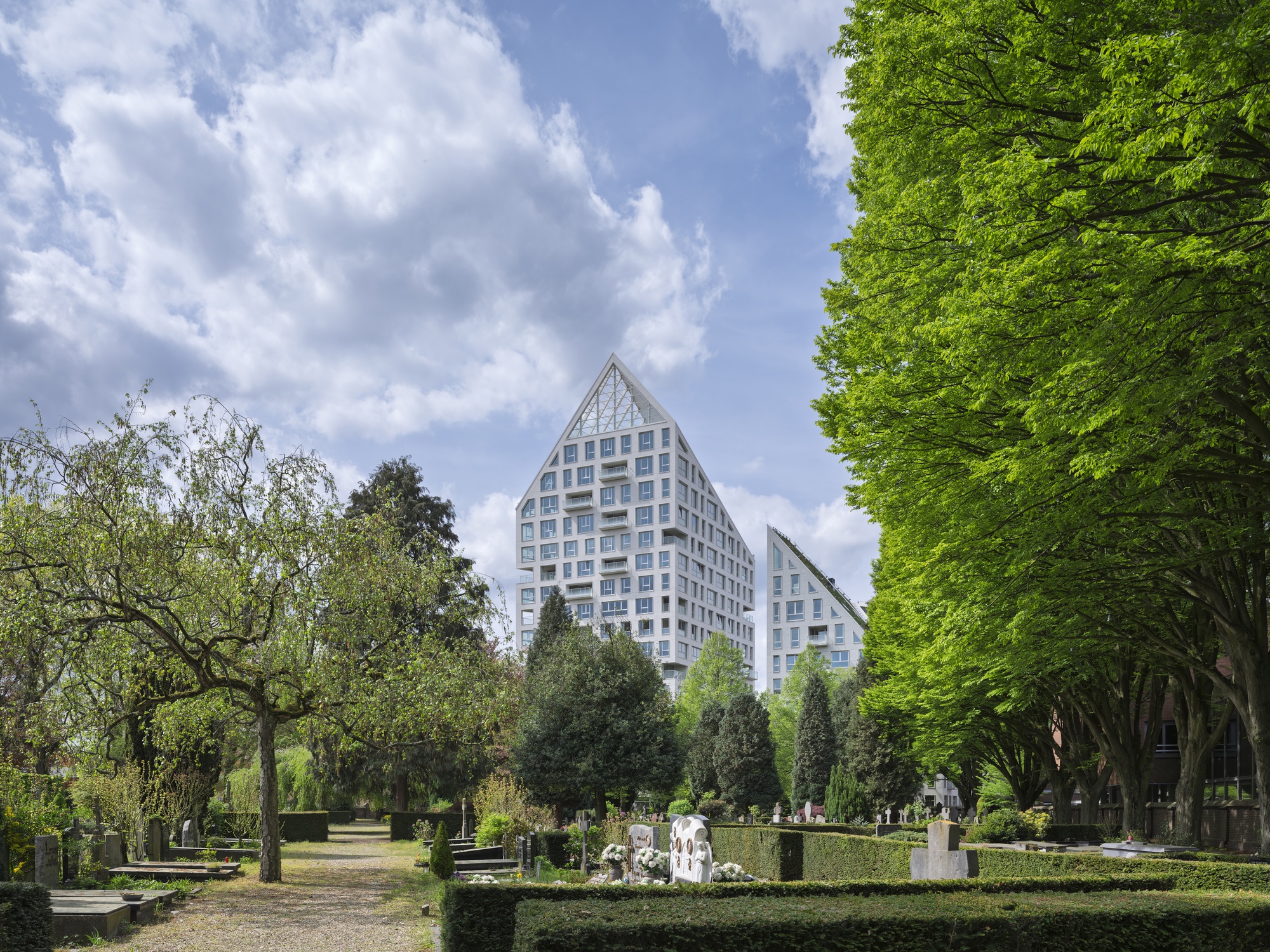 Twin gabled towers rising above a park cemetery with trimmed hedges and spring foliage