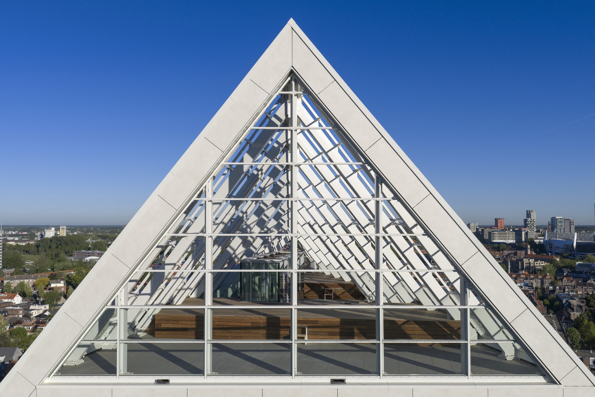 Triangular glass gable with exposed white structural frame against a clear blue sky and urban horizon