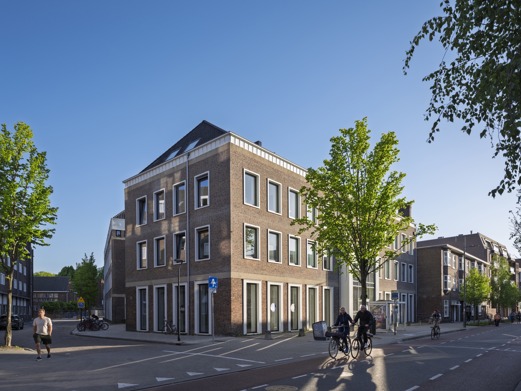 Street corner view of brick volume with white trim and planted trees with cyclists passing