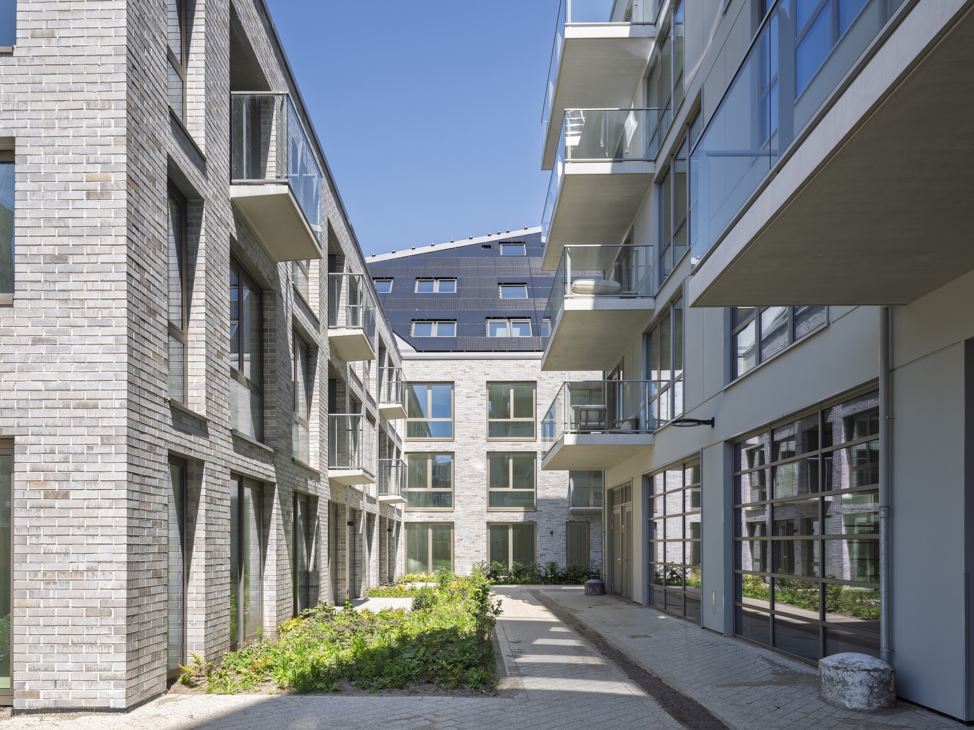 Courtyard passageway between brick facades with projecting balconies and planted beds in bright sunlight