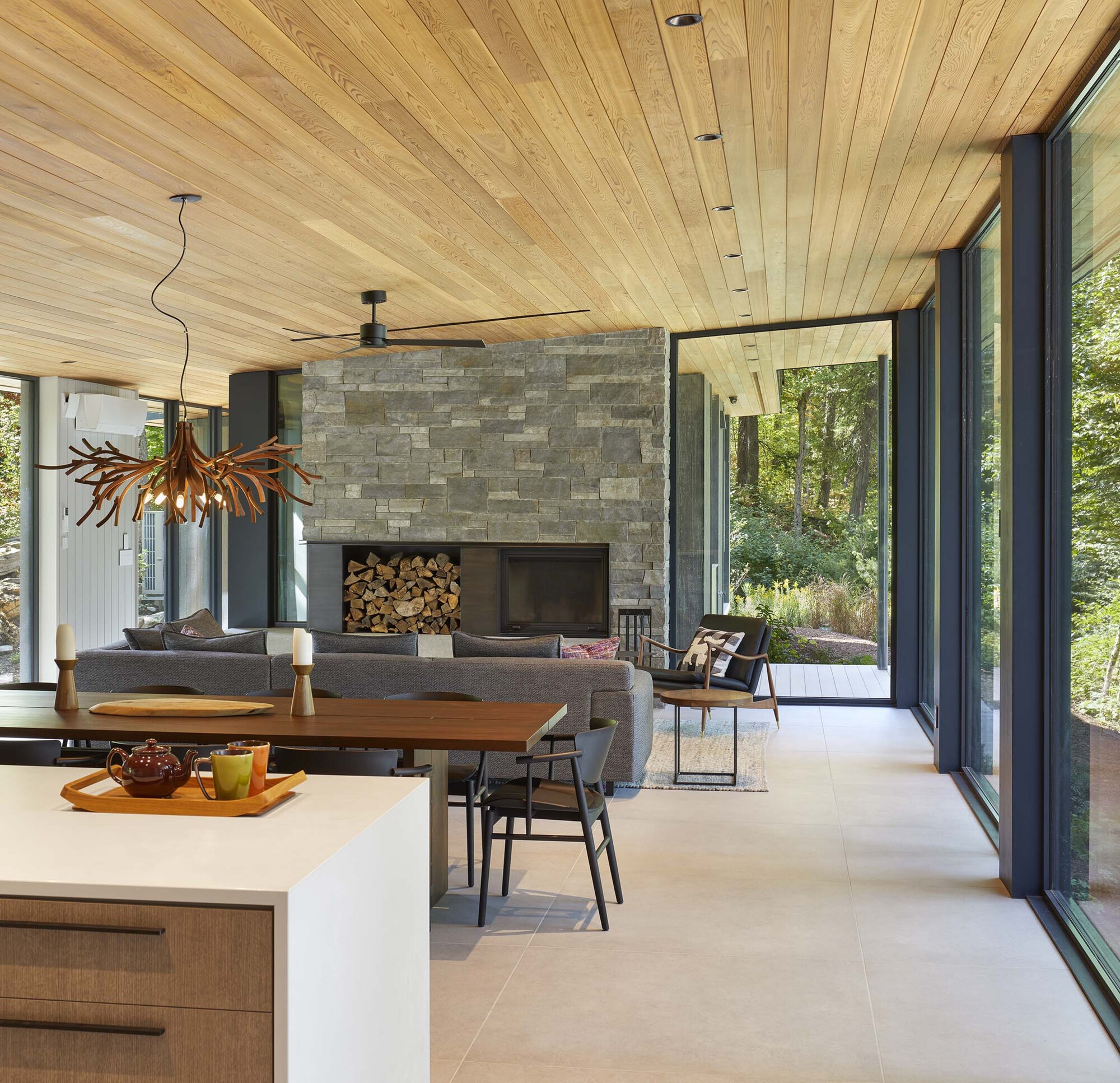 Open living space with stone fireplace, timber ceiling, and floor-to-ceiling windows framing woodland views
