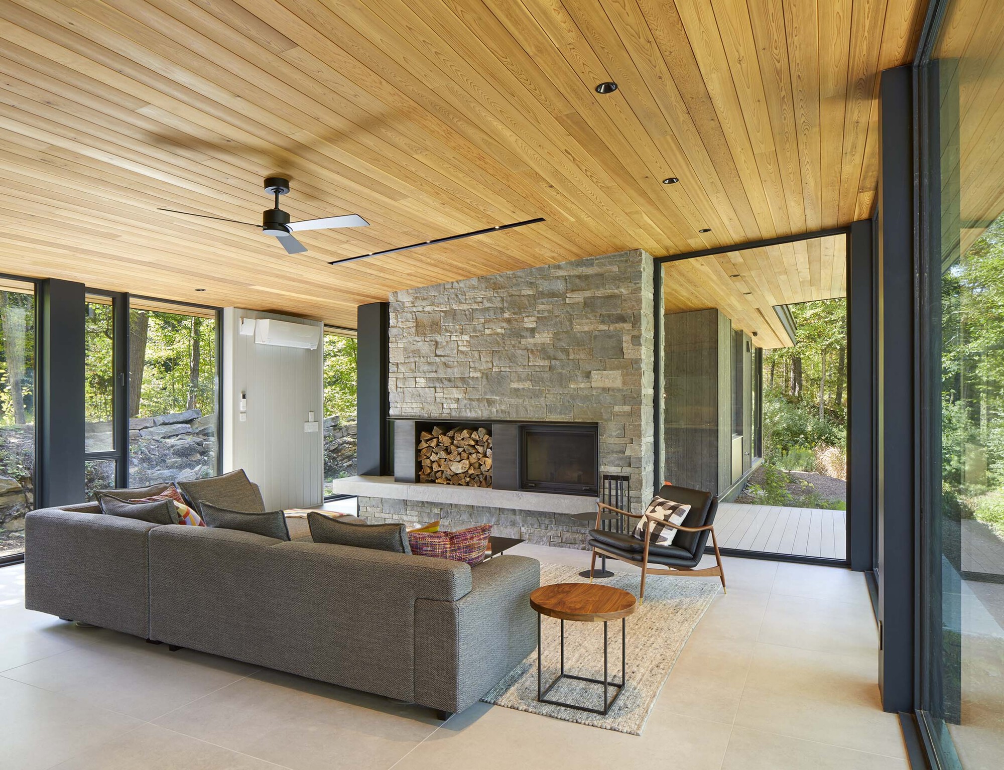 Living room with stacked stone fireplace, wood nook, and large glazed openings on two sides