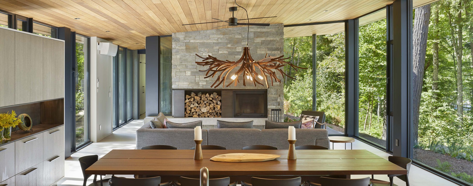 Open living and dining space with timber ceiling and stone fireplace framed by forest views