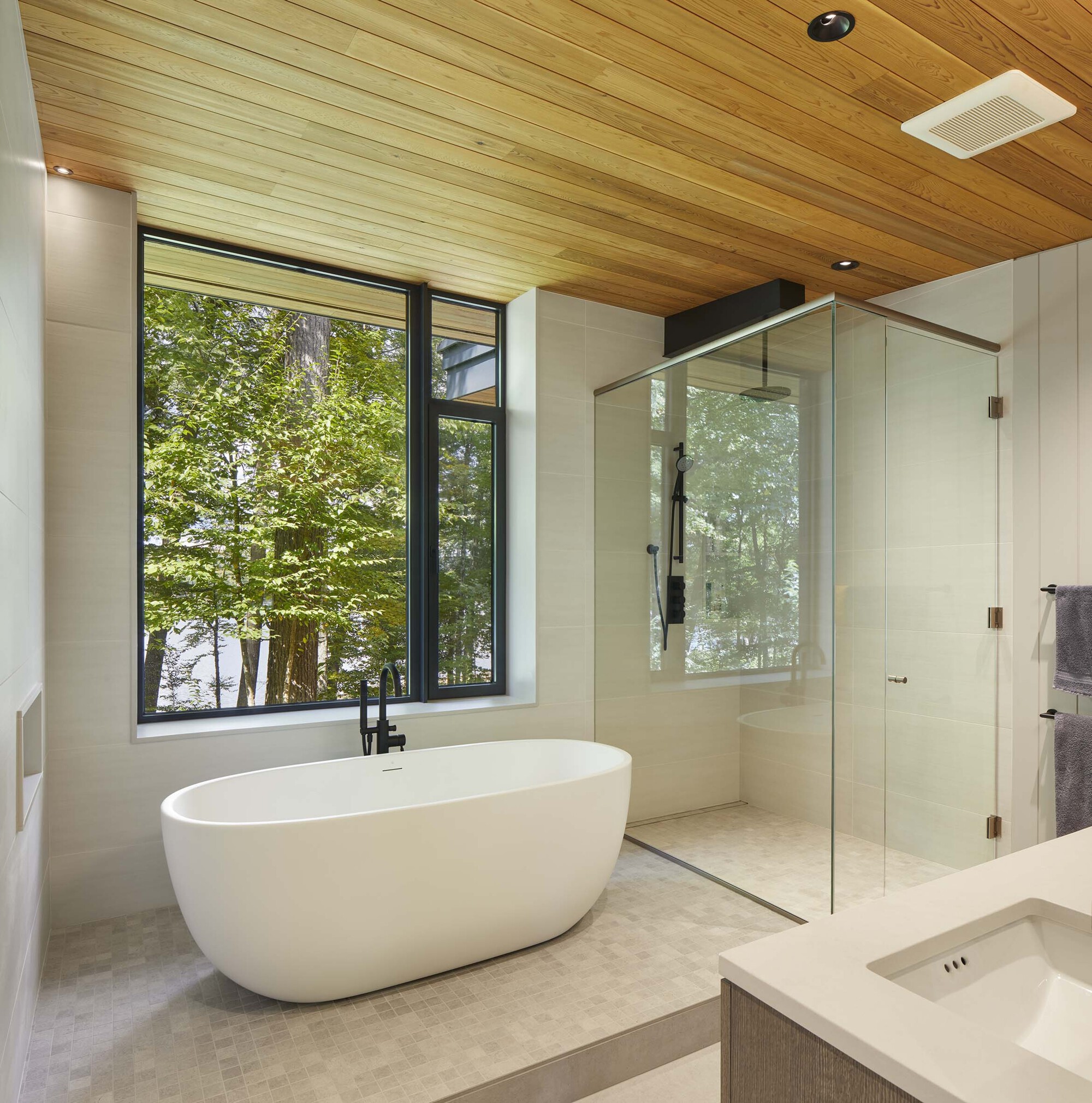 Bathroom with freestanding tub beneath timber slat ceiling and floor-to-ceiling window overlooking trees