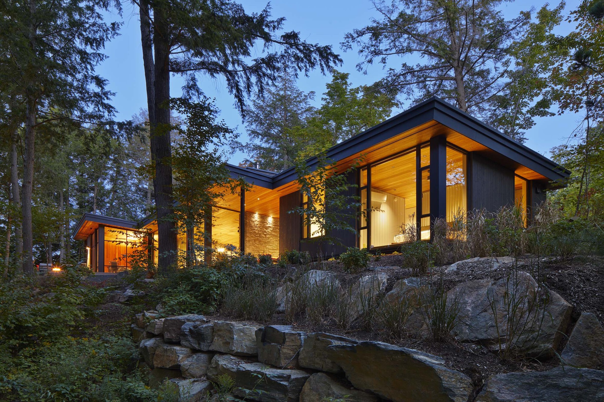 Illuminated residence with sloping roofline among boulders and native planting at twilight