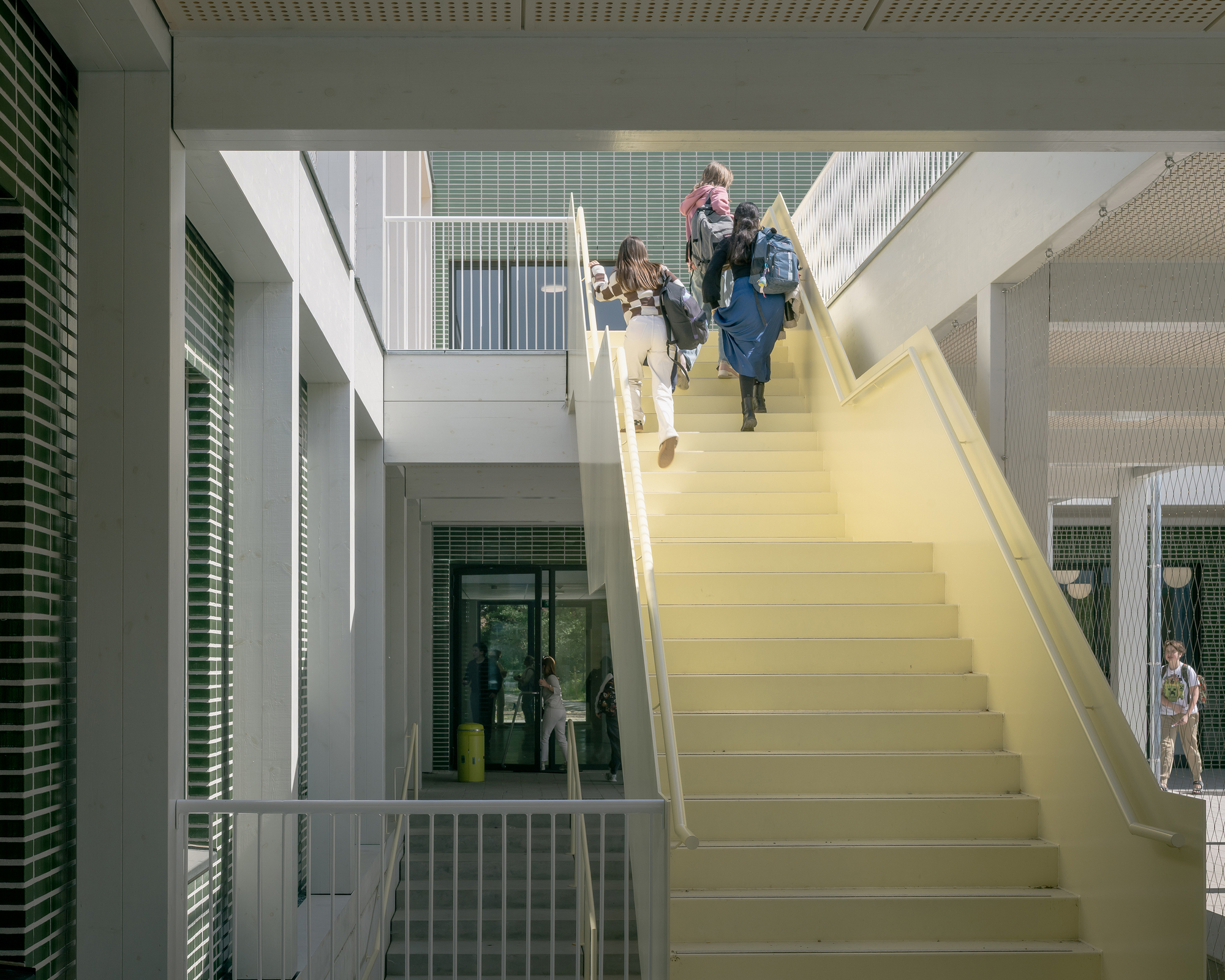 Yellow staircase ascending through a white atrium with green louvered screens and students climbing between levels