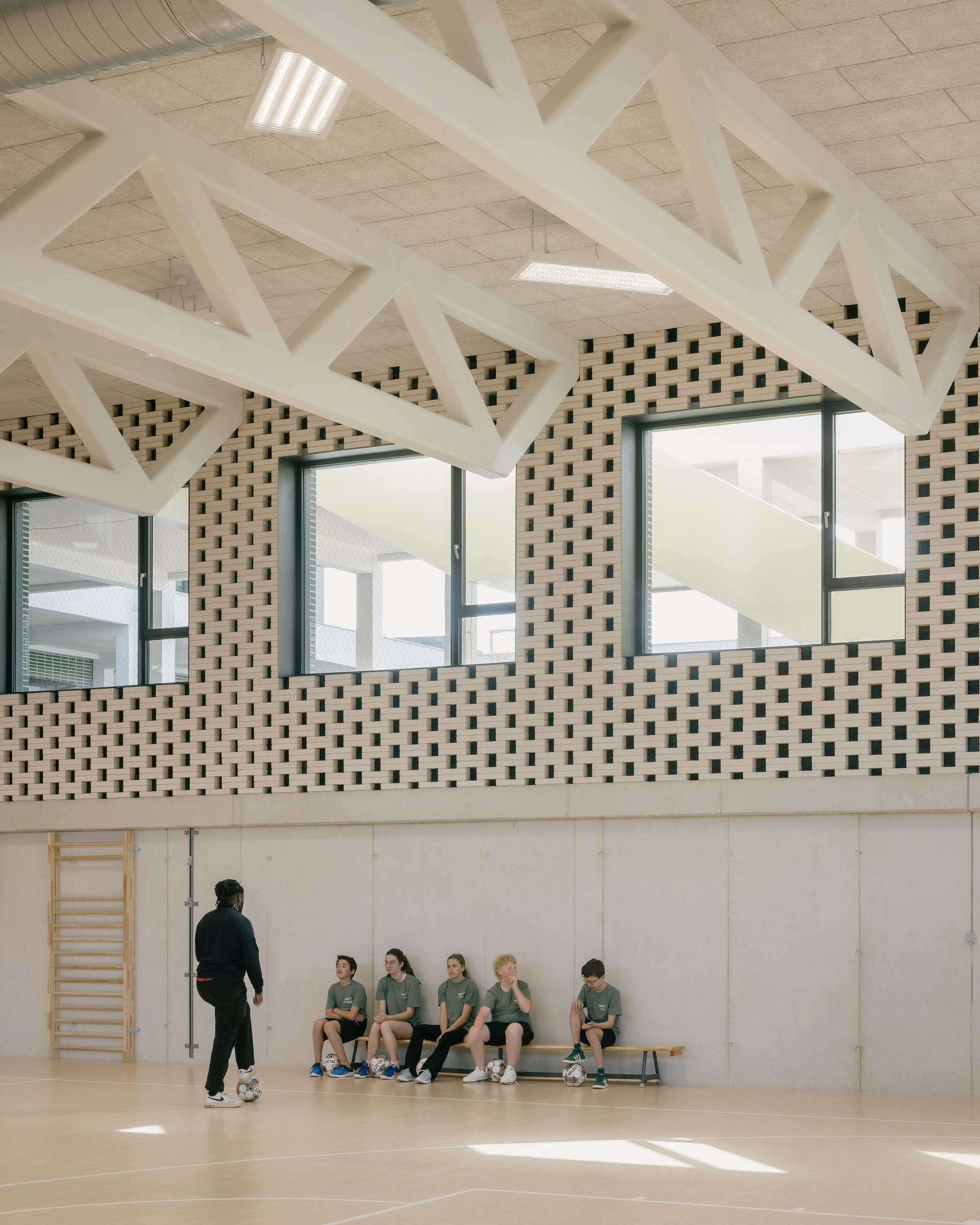Gymnasium with chequered brick clerestory wall and white timber trusses above children seated on a bench