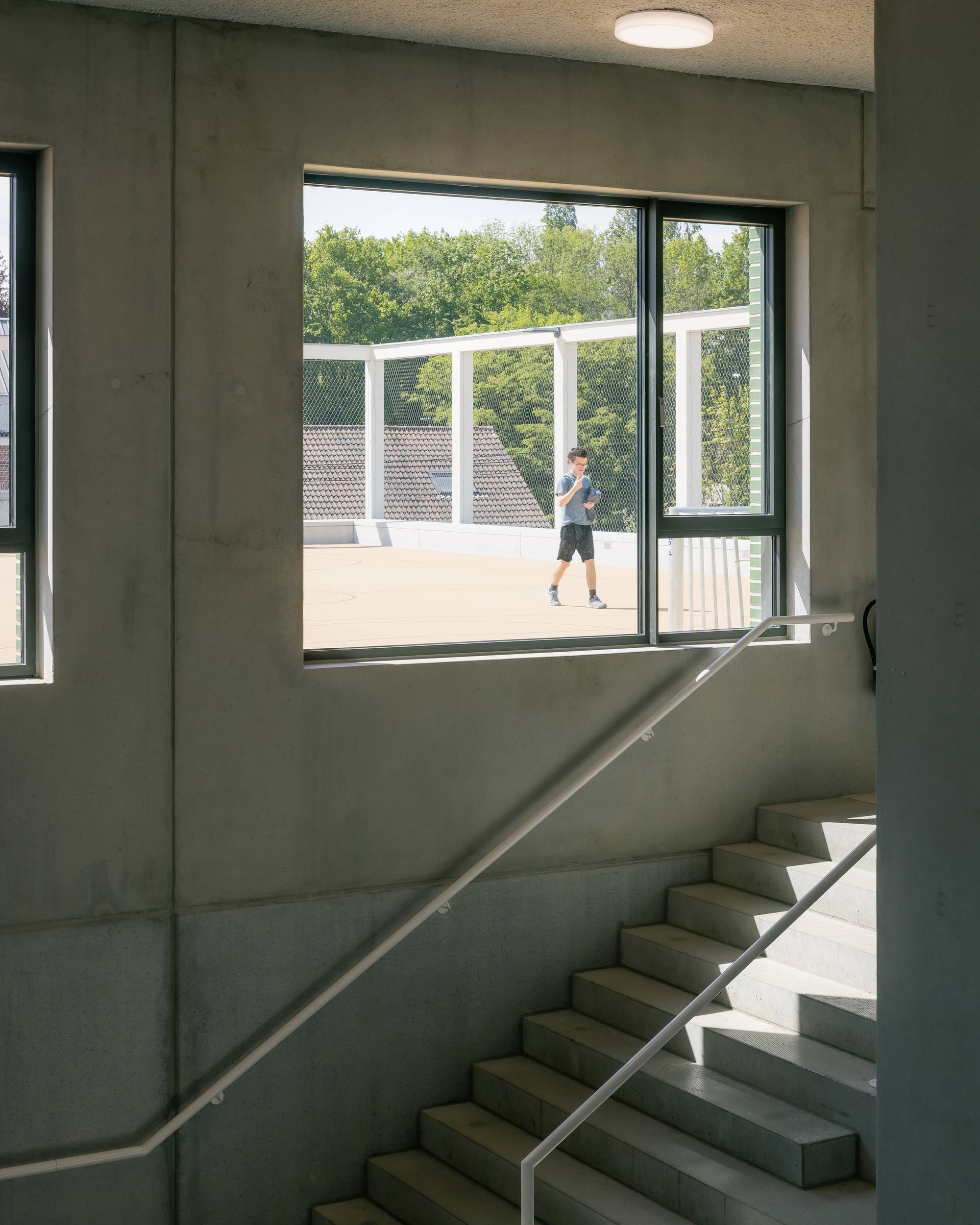 Concrete stairwell with steel handrail framing a view to an outdoor court where a student walks in sunlight