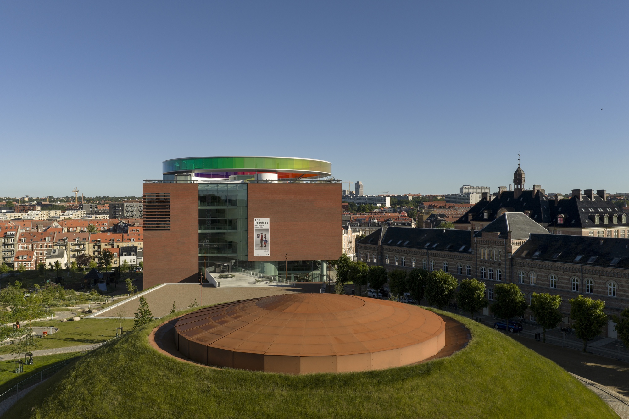 Cylindrical building with terracotta panels and rainbow rooftop overlooking the surrounding urban district