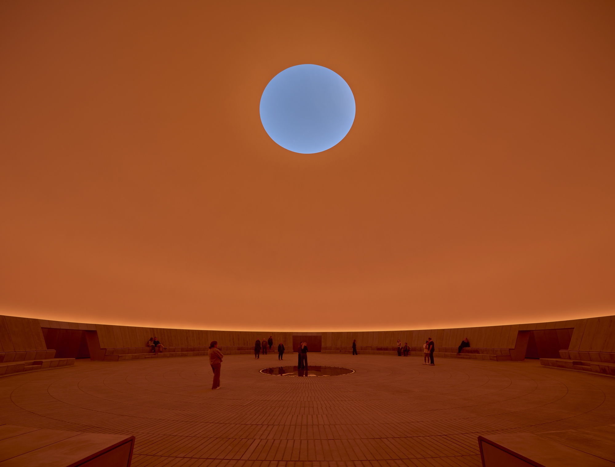 The same circular courtyard space illuminated with warm orange light and visitors on the floor