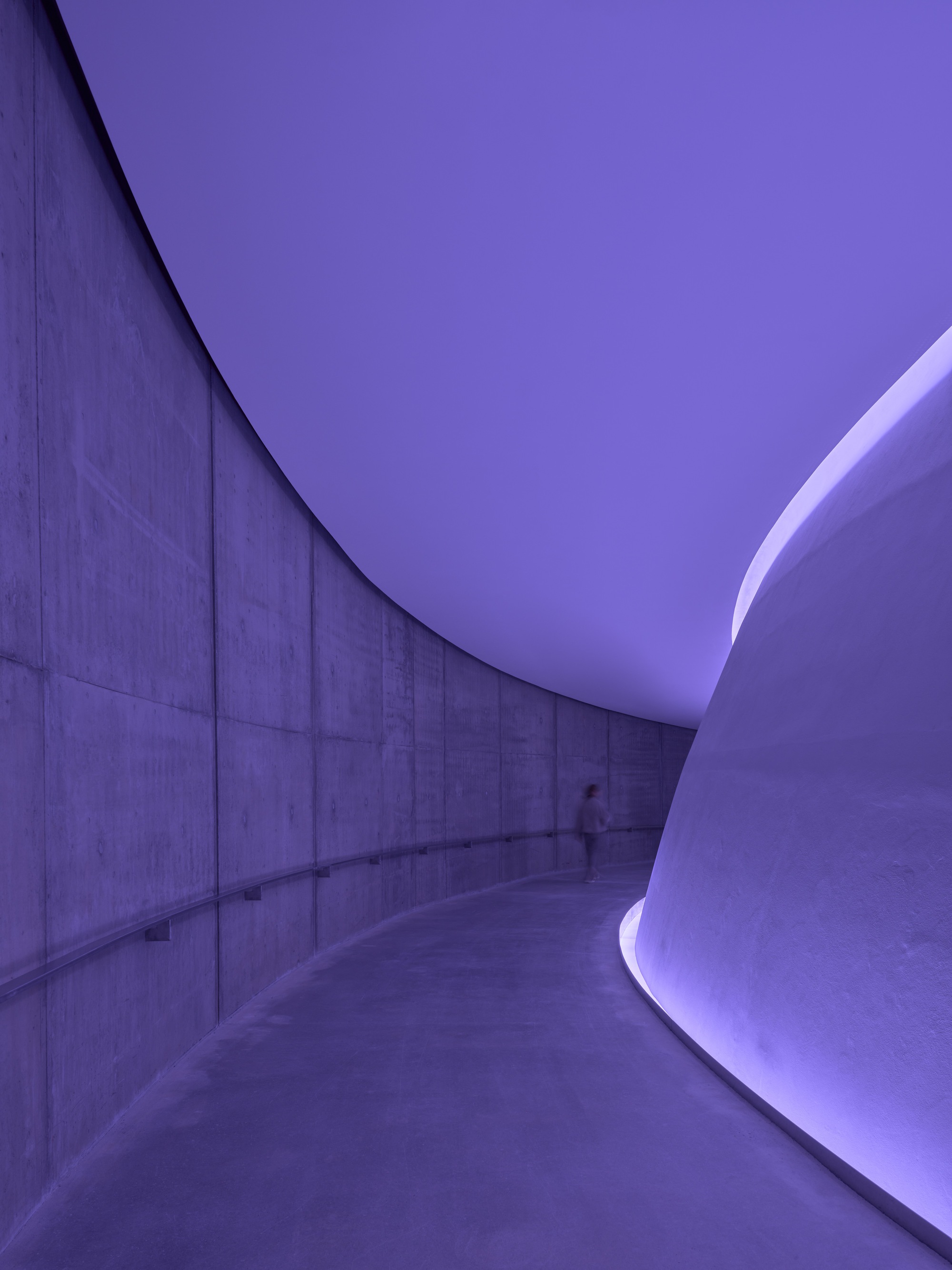Curved concrete corridor with purple light washing the ceiling and a solitary figure walking