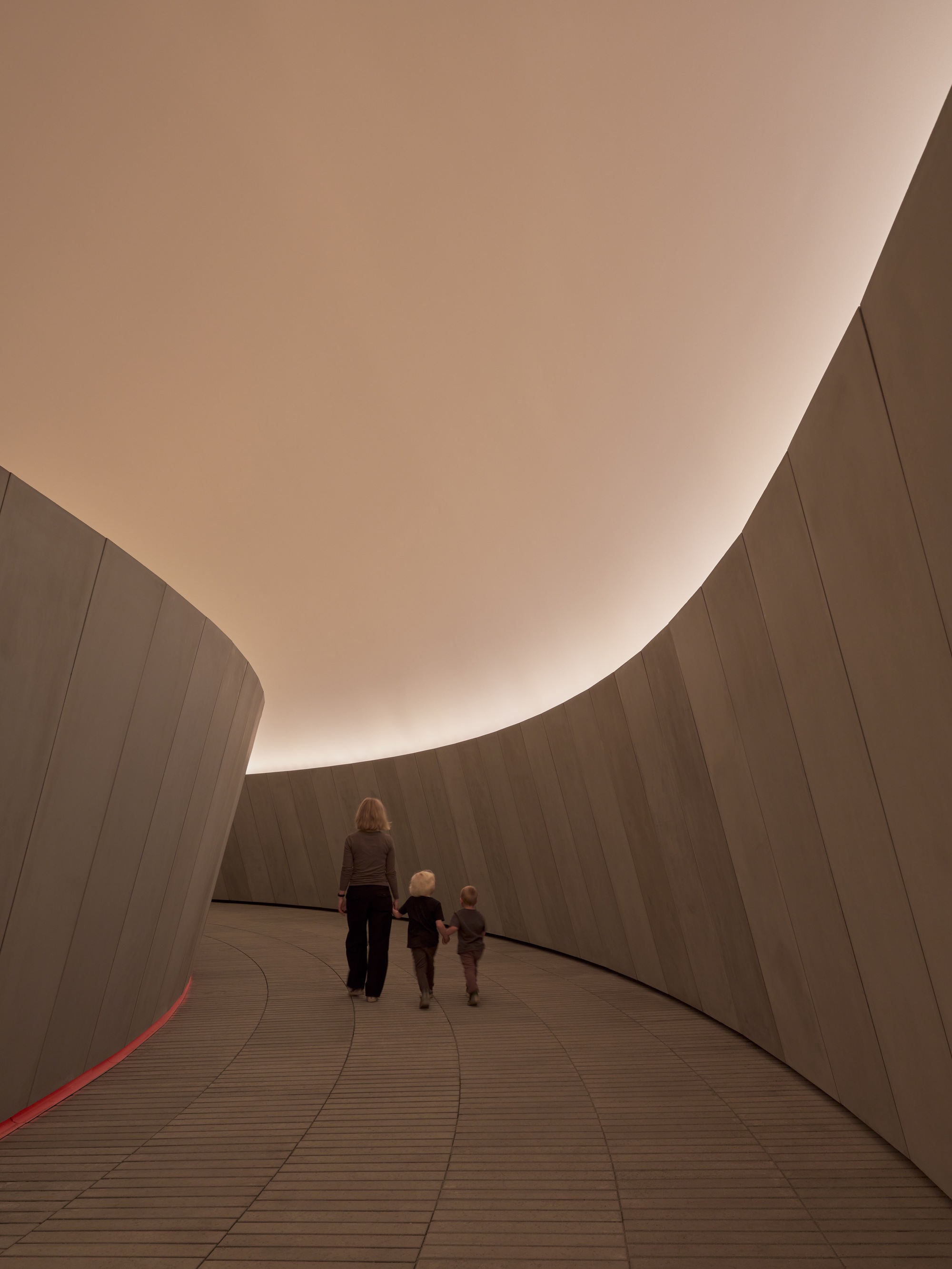 Ramped passageway with concrete walls and warm overhead light as visitors ascend together