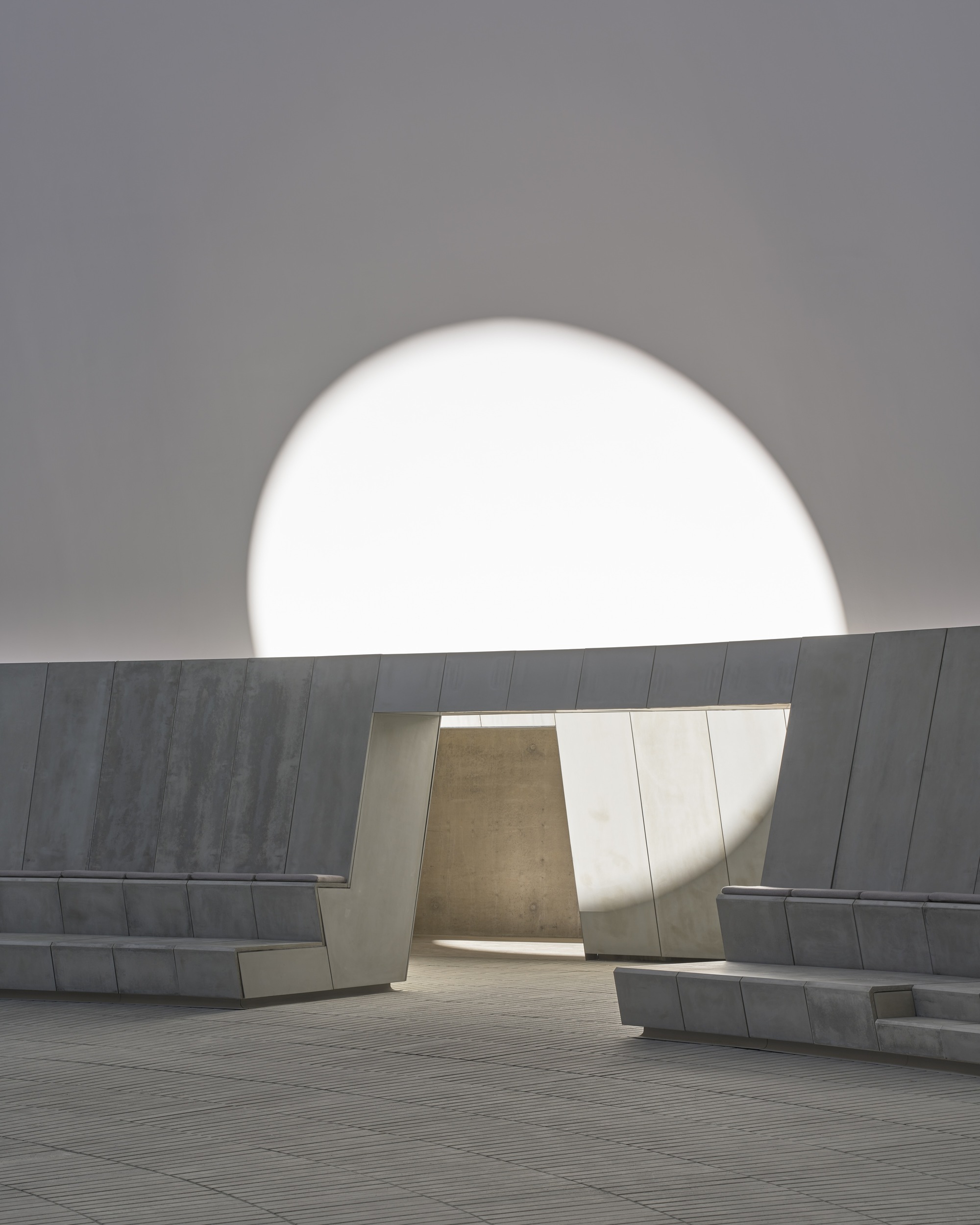 Courtyard portal framing a distant stone volume beneath a monumental circular oculus in the sky