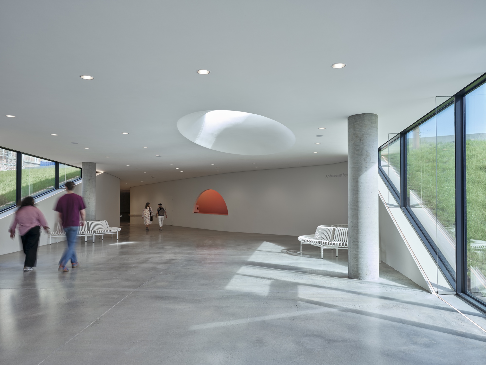 Interior foyer with circular ceiling aperture, concrete column, and visitors moving through sunlit glazed corridors