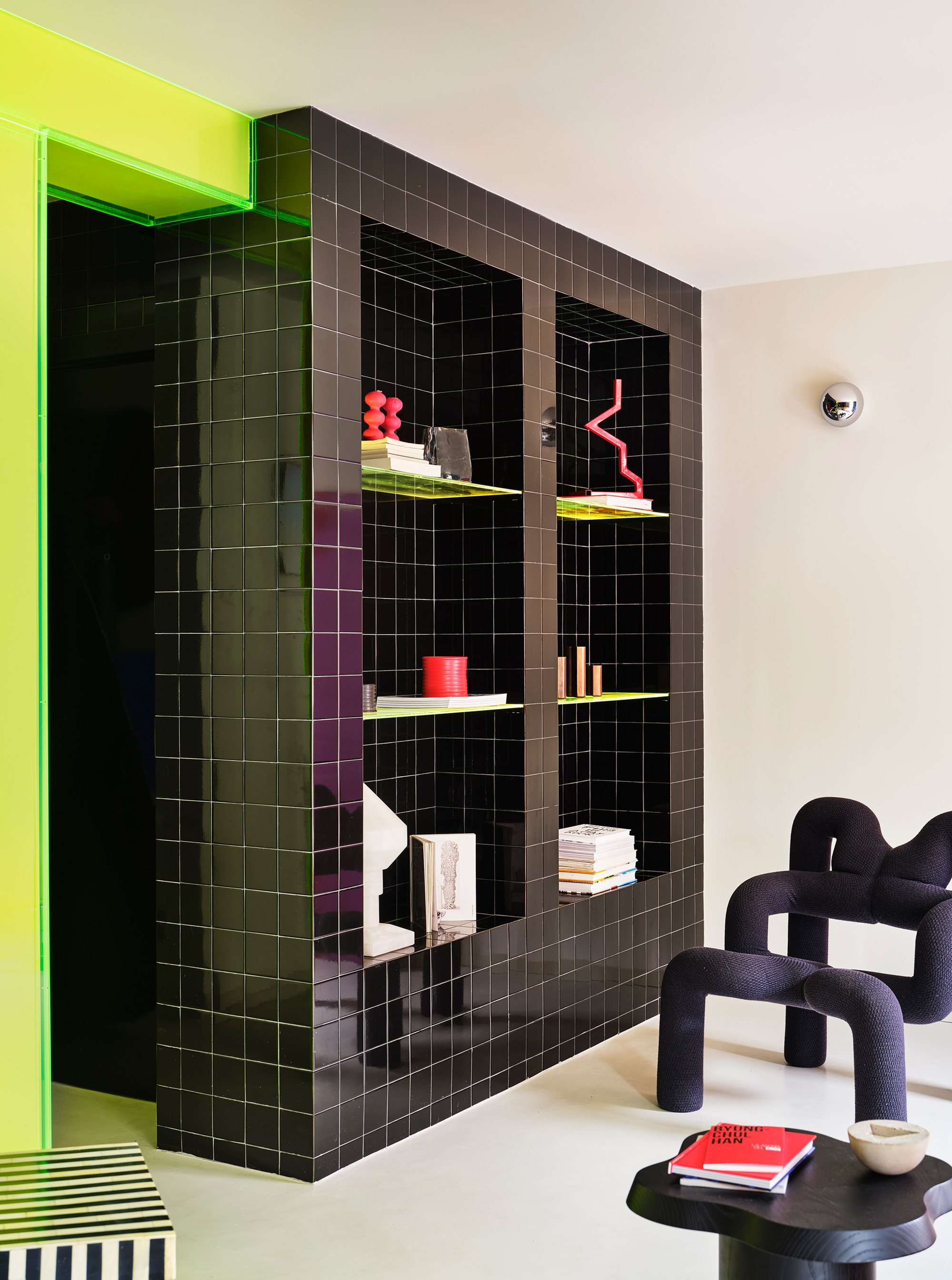 Black tile shelving unit with yellow acrylic shelves displaying objects against a chartreuse framed opening