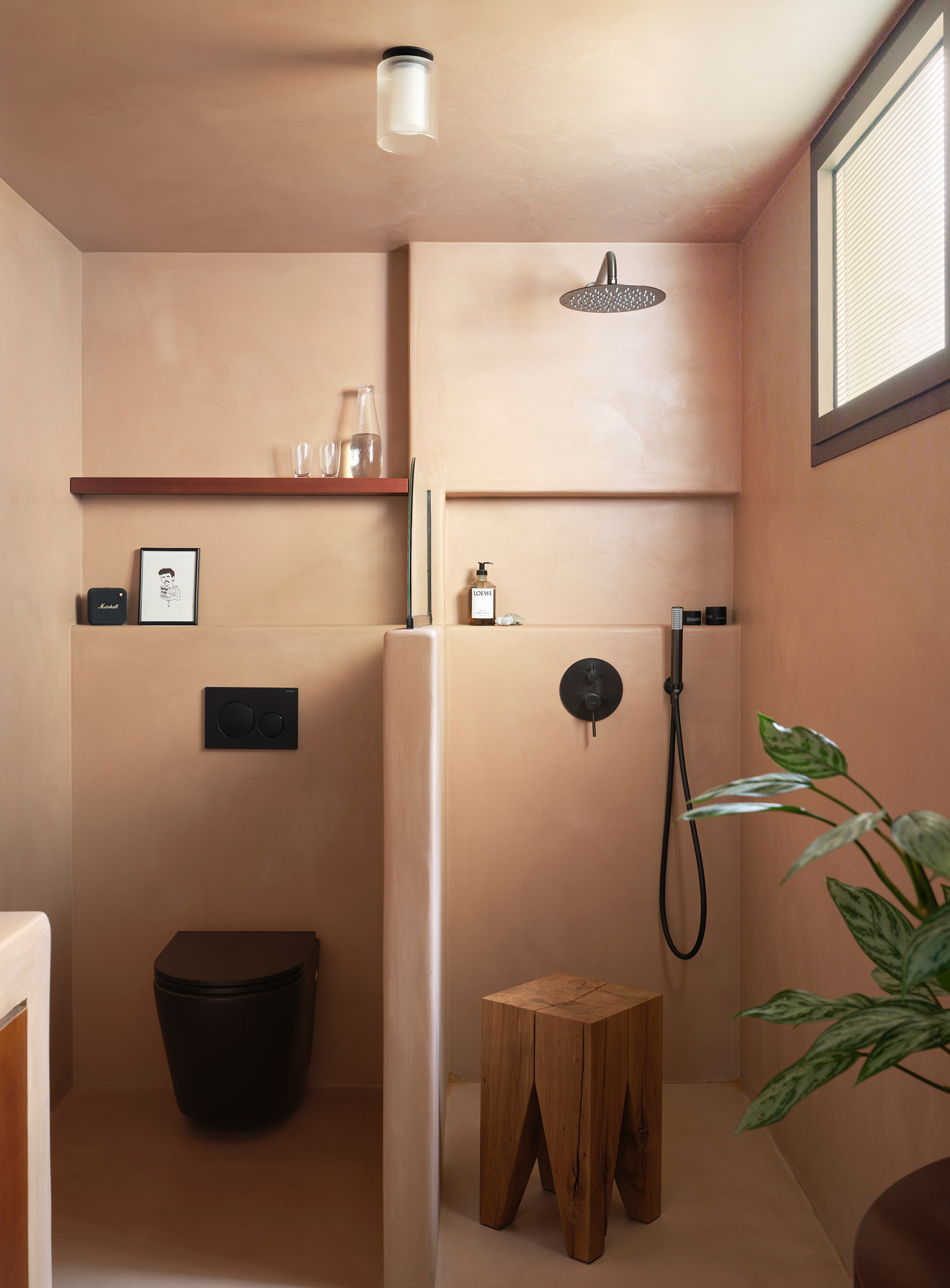 Bathroom interior with peach-toned plaster walls, black fixtures, timber shelf and stool, and potted plant