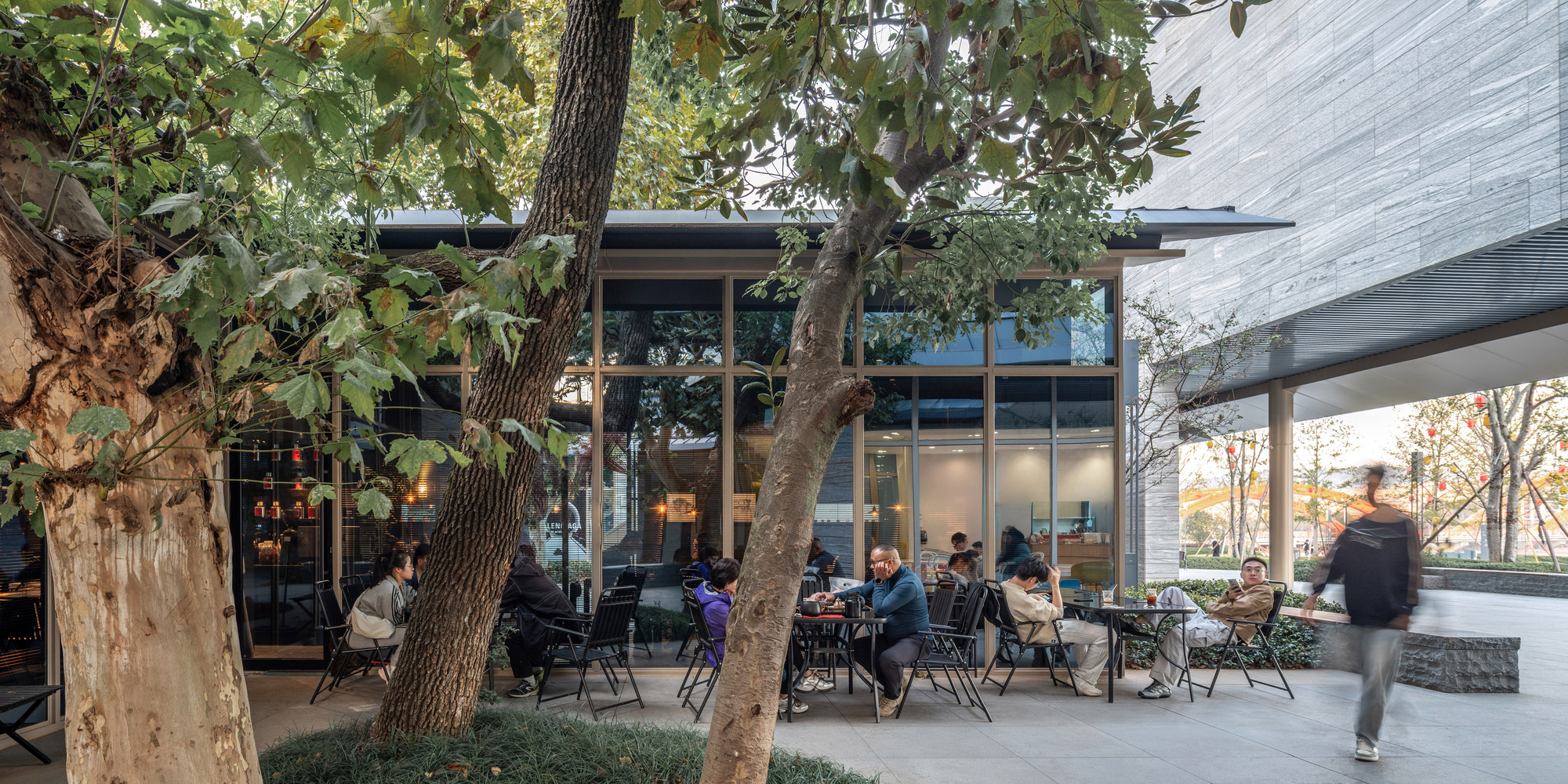 Outdoor cafe seating under a tree canopy with glass pavilion and passing pedestrian