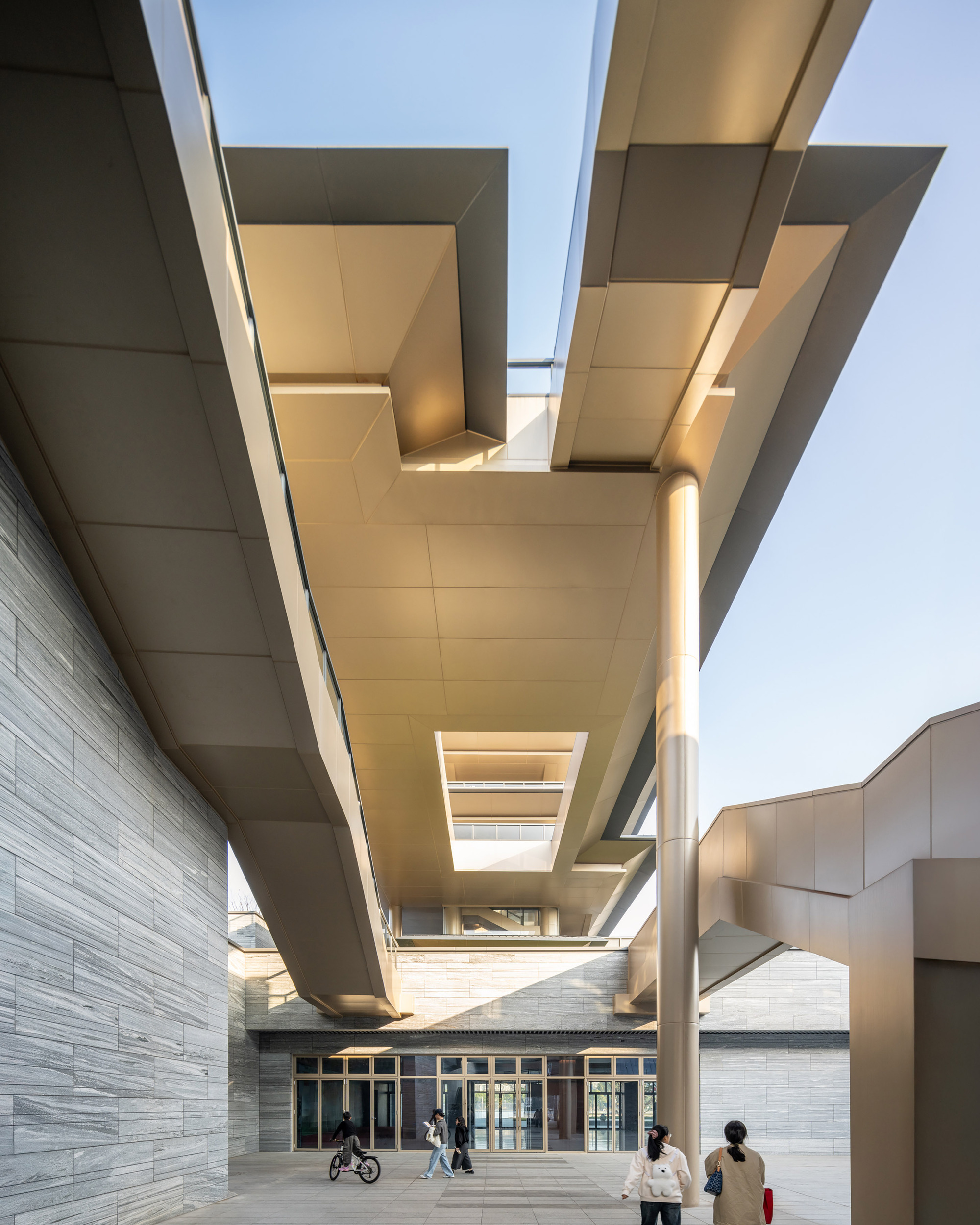 Entry courtyard beneath intersecting cantilevers with horizontal stone cladding and visitors walking below