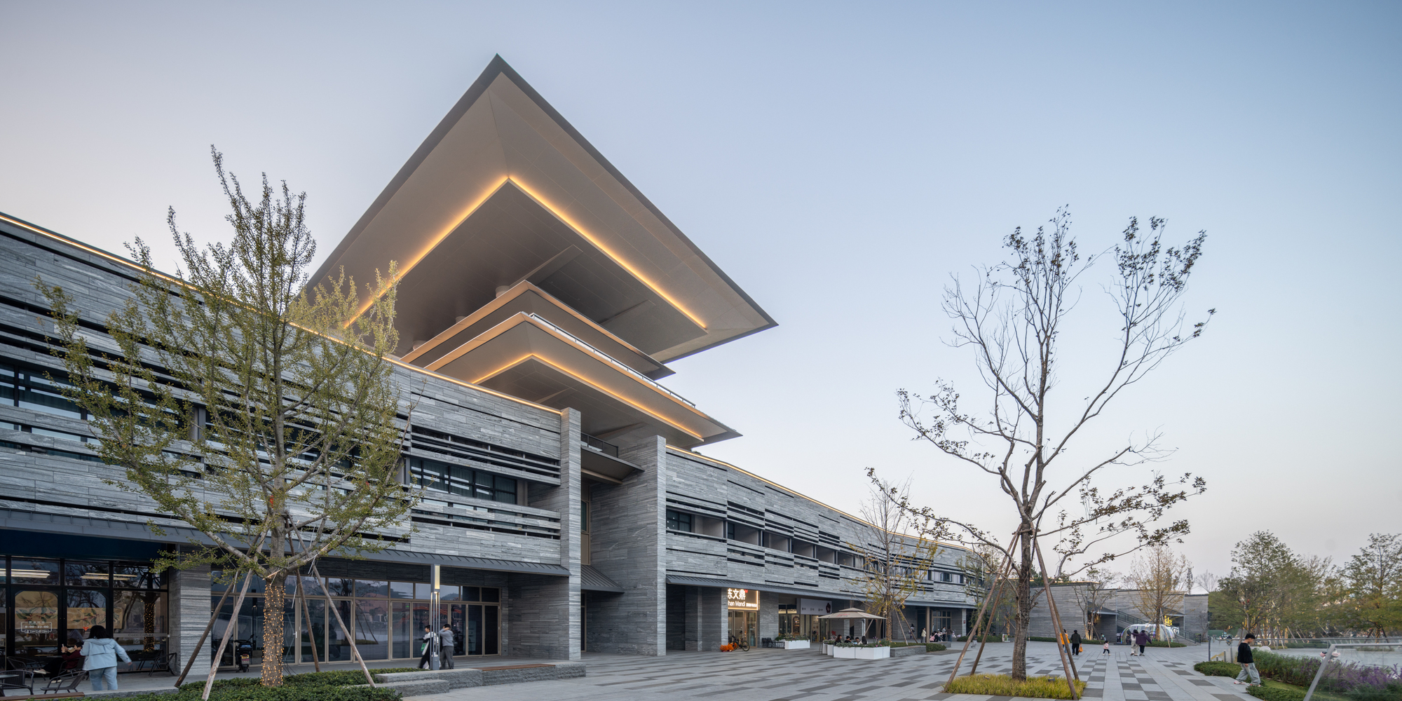 Long facade view showing the tiered illuminated canopy above horizontal louvers and young trees at dusk