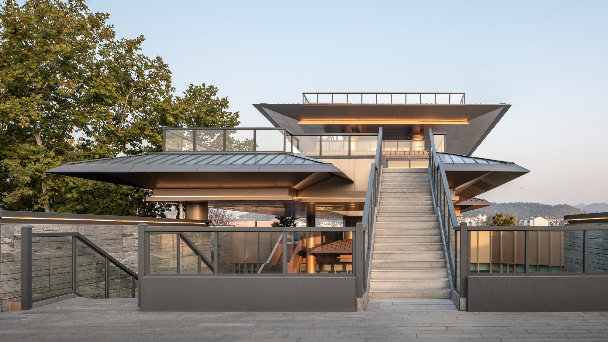 Exterior stair rising to the layered roof structure with glass railings and trees at twilight