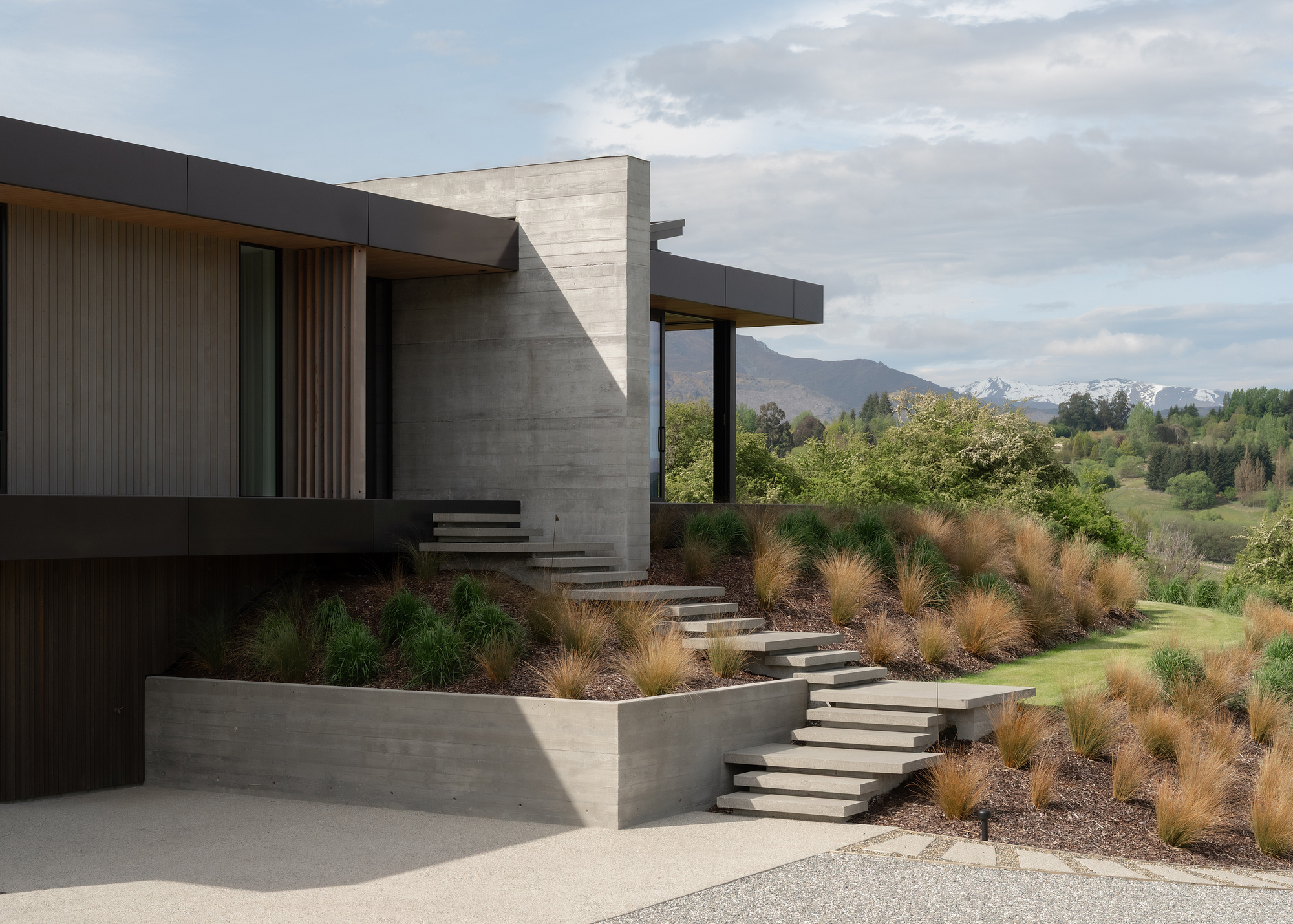 Concrete retaining walls and stepped pathway through native grasses leading to the entrance under cloudy skies