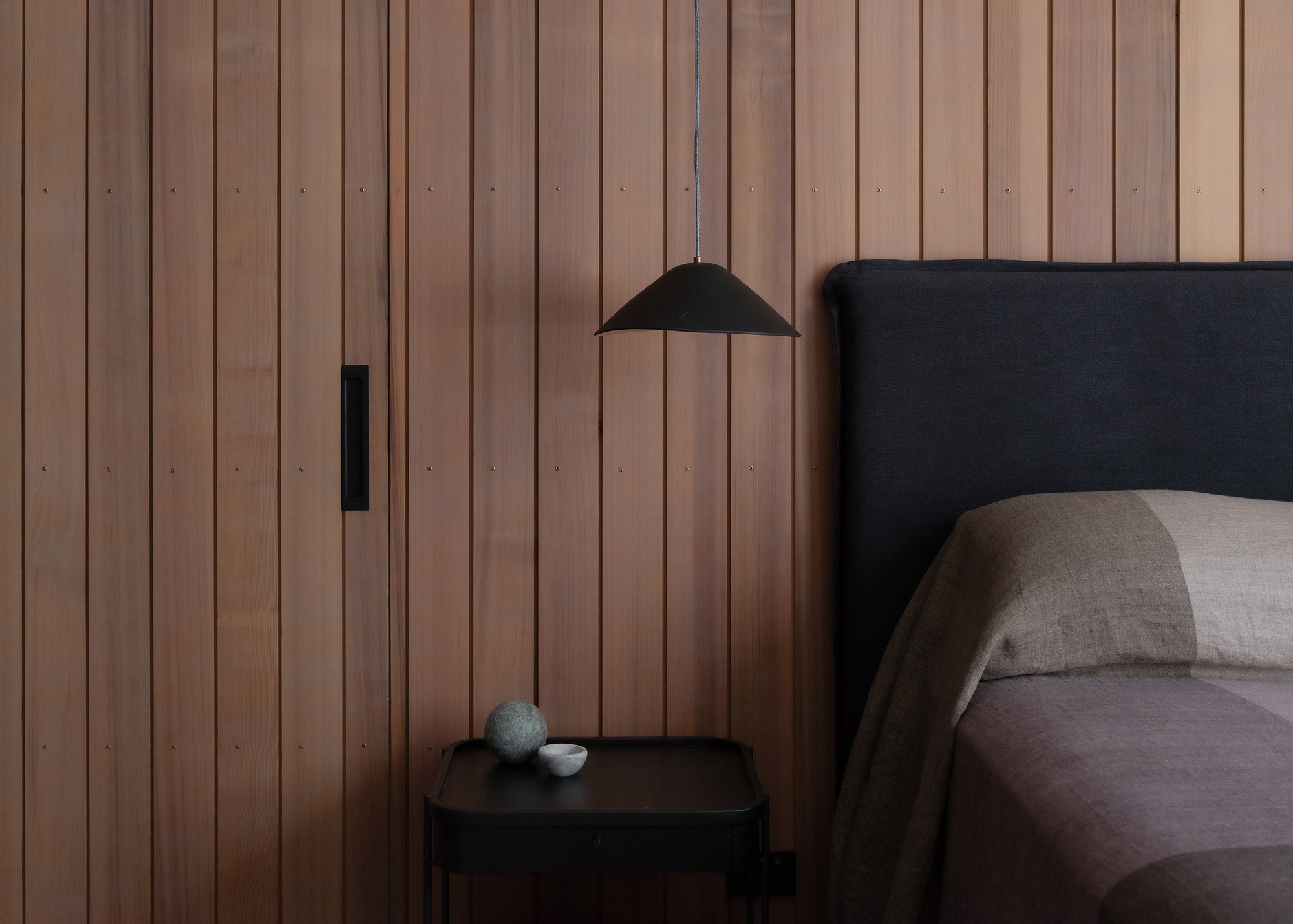 Bedroom detail showing vertical timber cladding, pendant light and upholstered headboard beside a nightstand