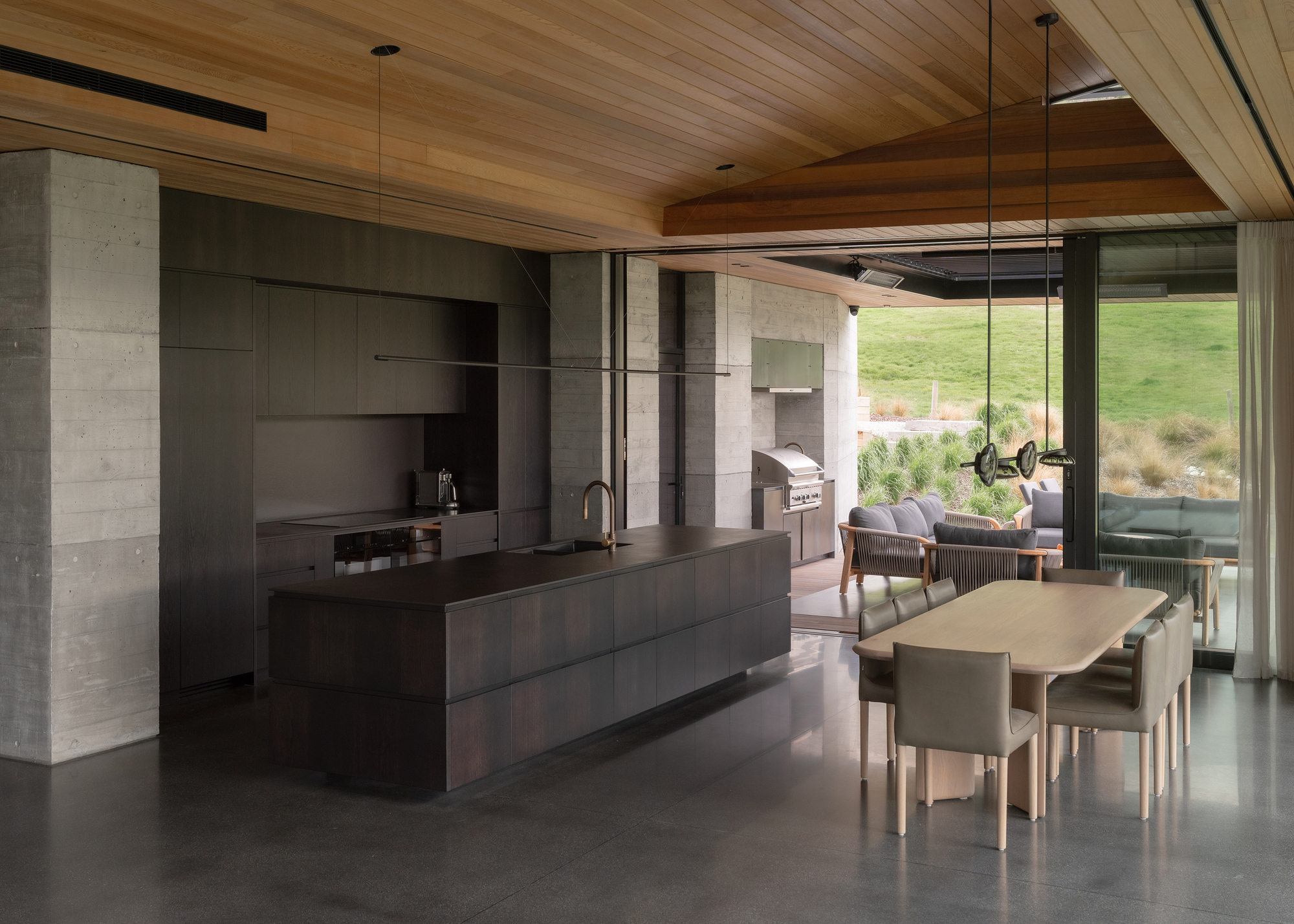 Open kitchen and dining area with dark timber island, wood-slat ceiling and glazed doors to landscape