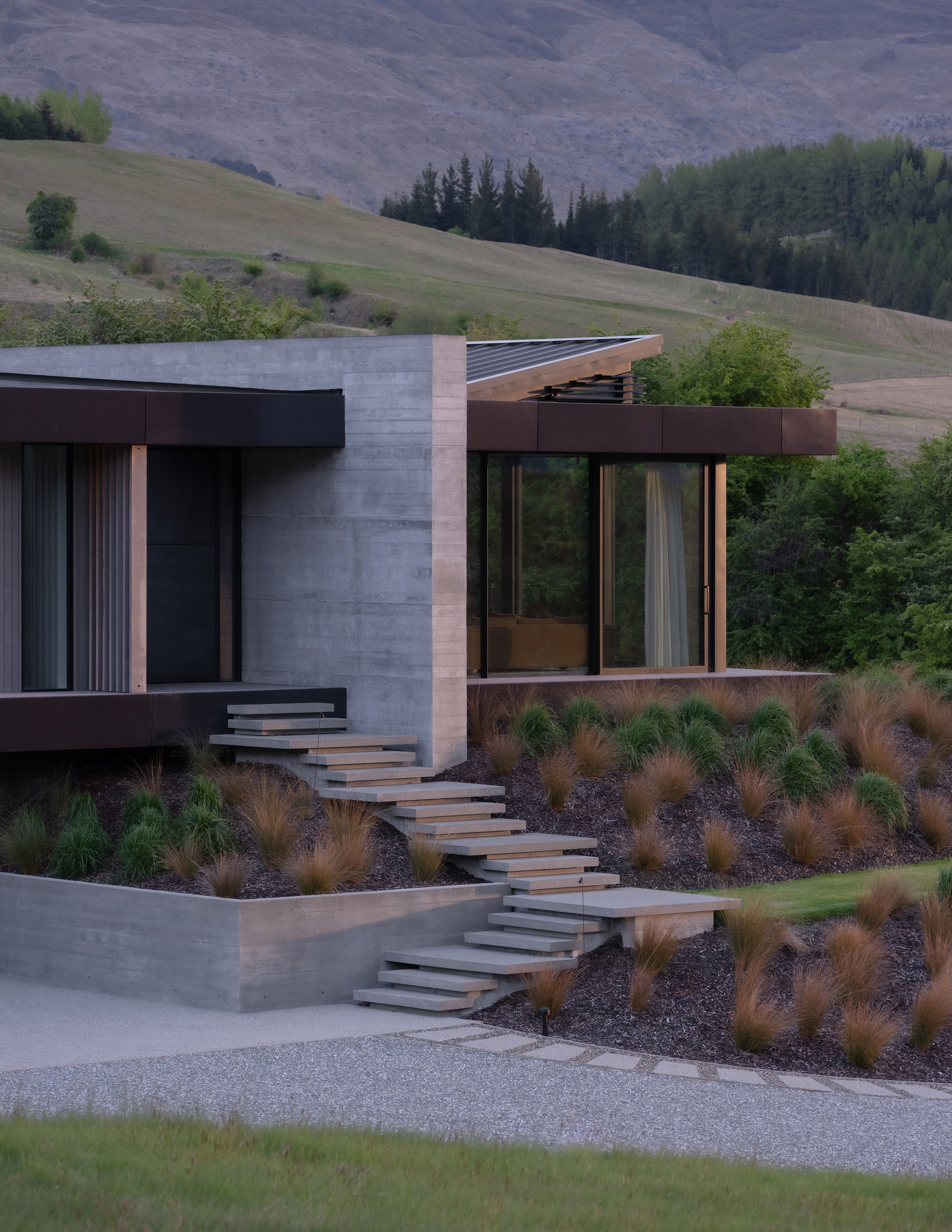 Concrete and timber facade with floating entry stairs through planted hillside garden at dusk