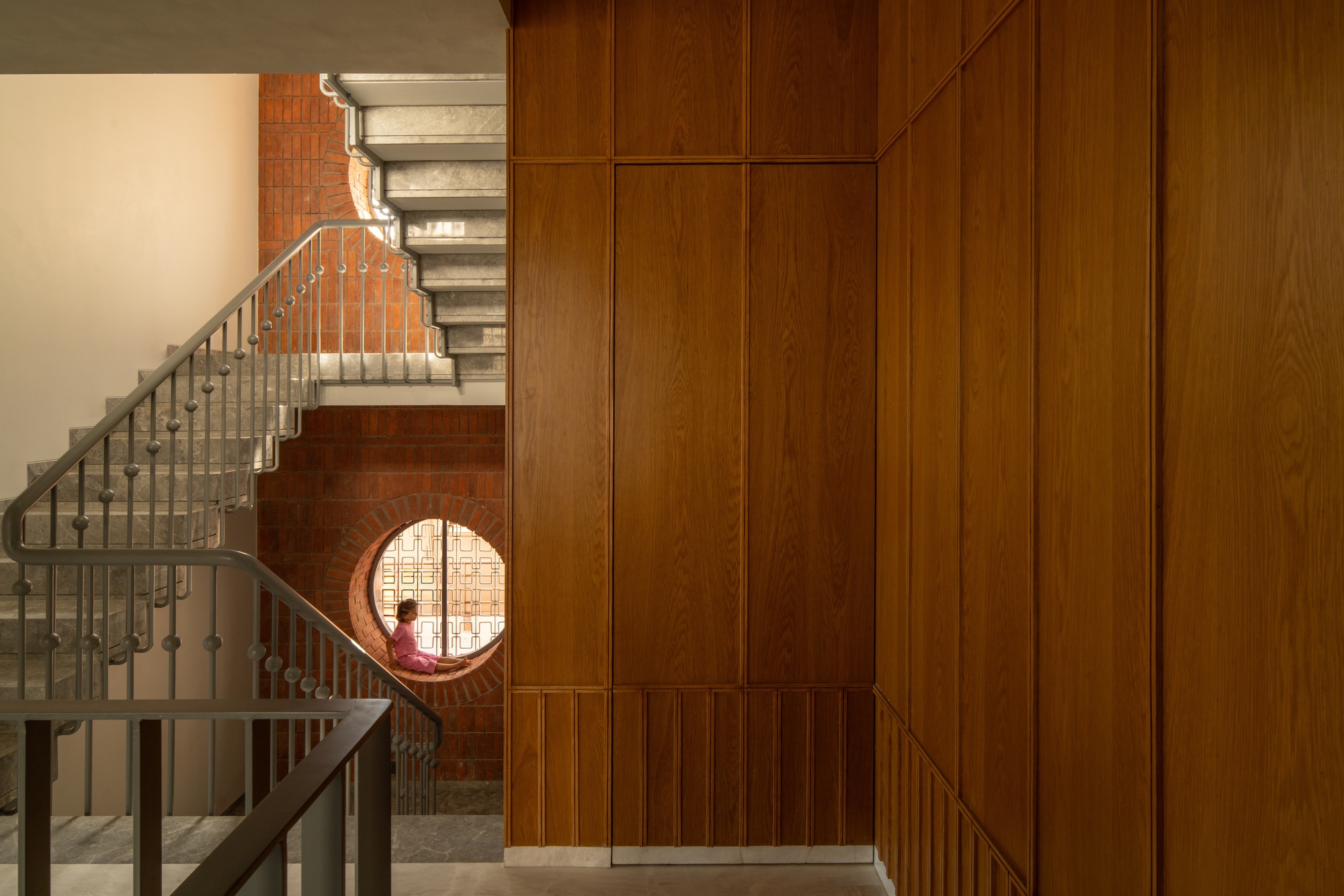 Concrete staircase with metal railing ascending alongside timber-clad walls and a circular brick-framed window with seated figure