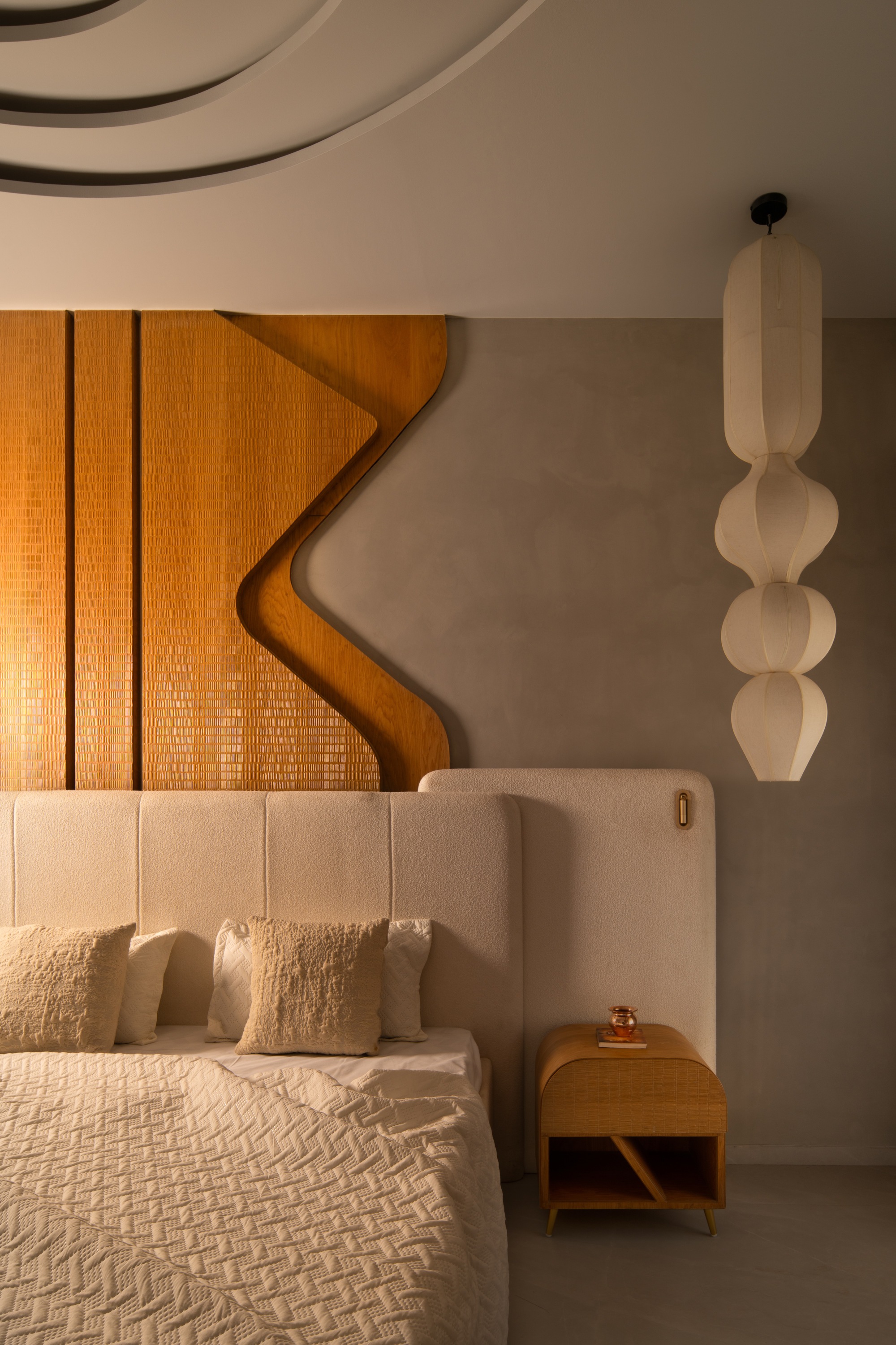 Detail of the upholstered bed headboard with curved timber panel above and pendant light hanging nearby
