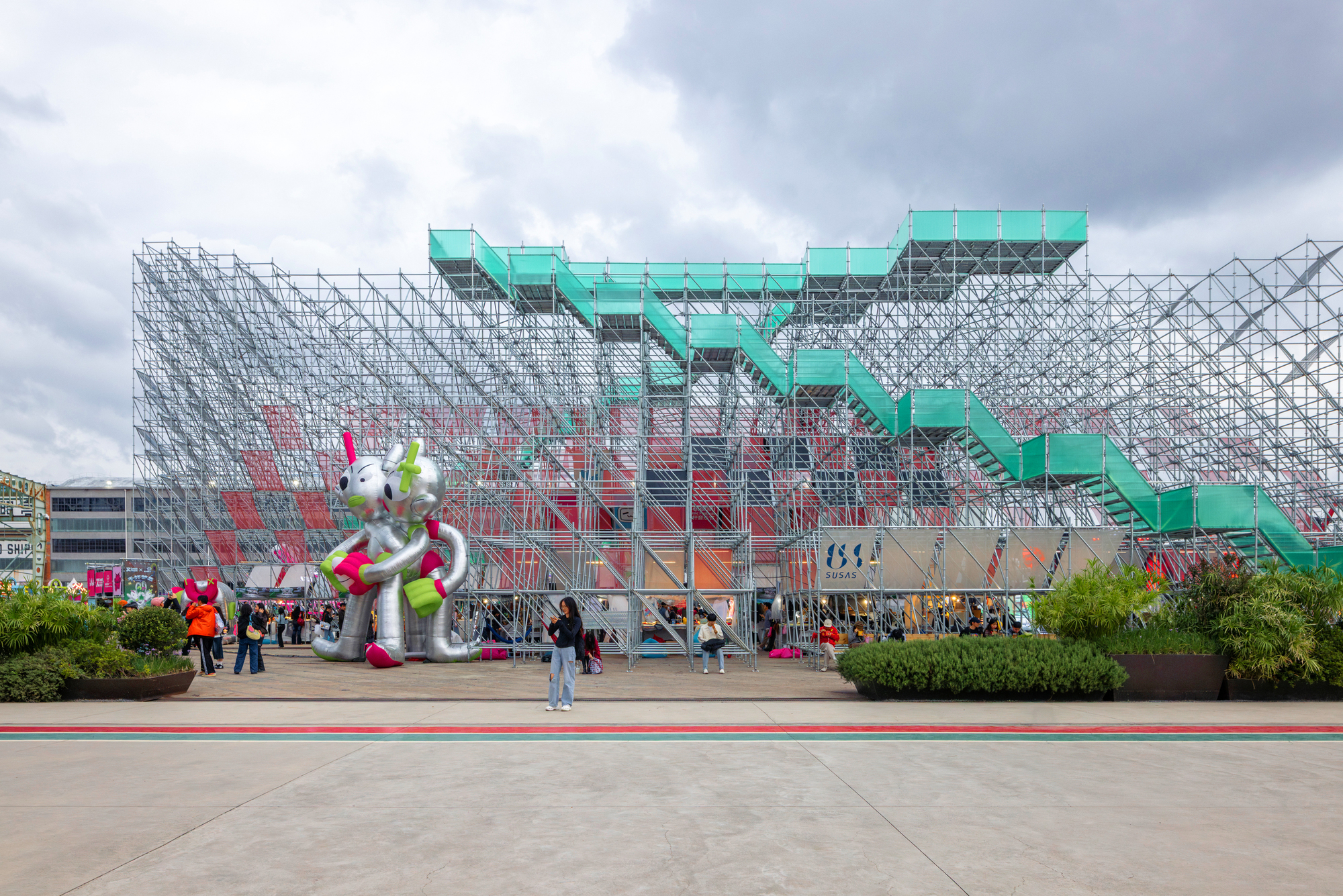Tubular steel scaffolding structure with turquoise stairs and visitors on the plaza under cloudy skies