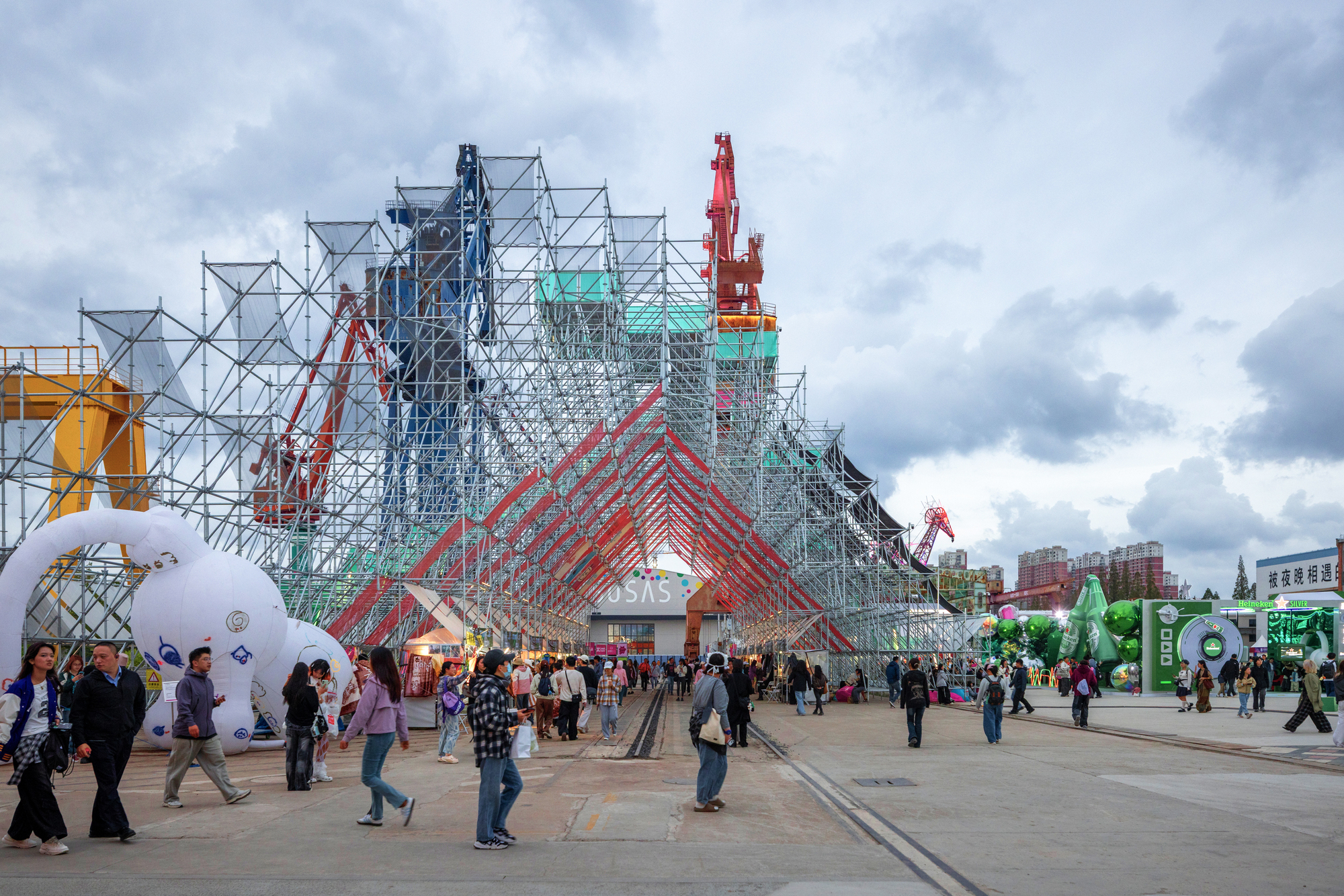 Gabled scaffolding entrance with red metal framework and crowds gathering beneath overcast afternoon light