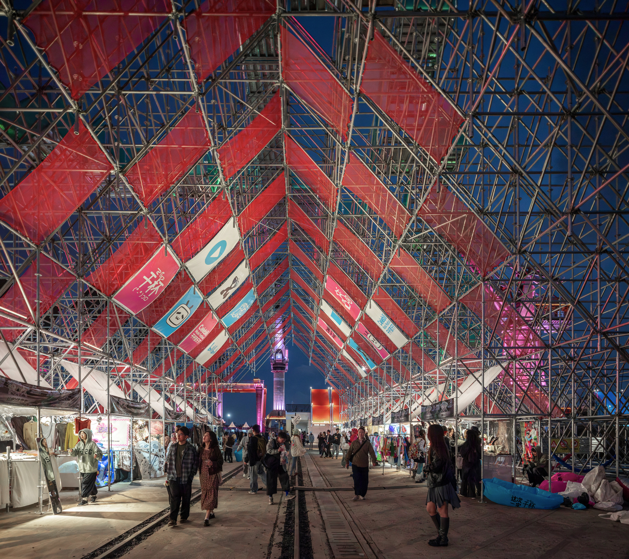 Interior passageway under the triangular metal scaffolding canopy with hanging banners and evening visitors