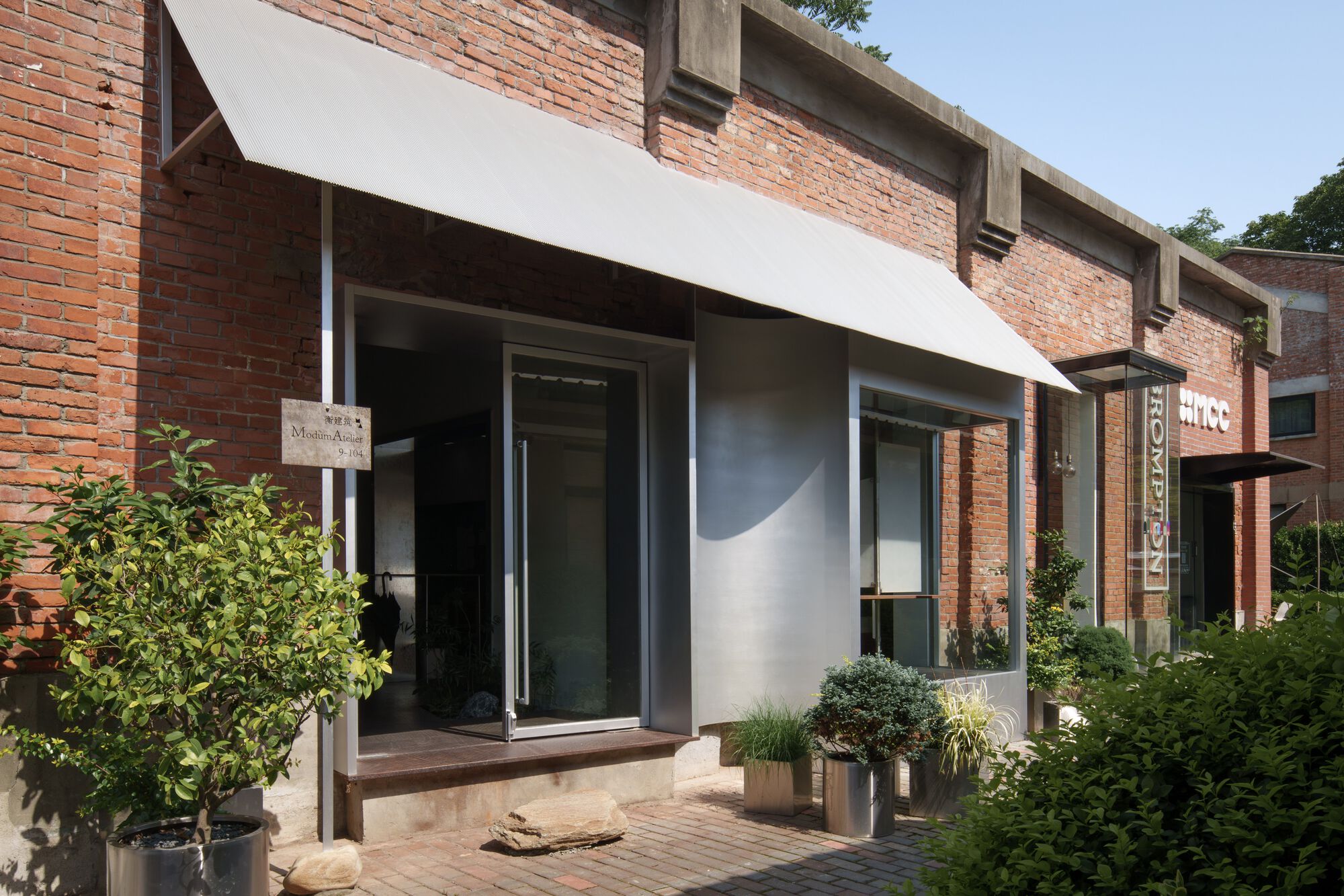 White-framed entrance set within an existing brick facade with potted plants on a paved courtyard