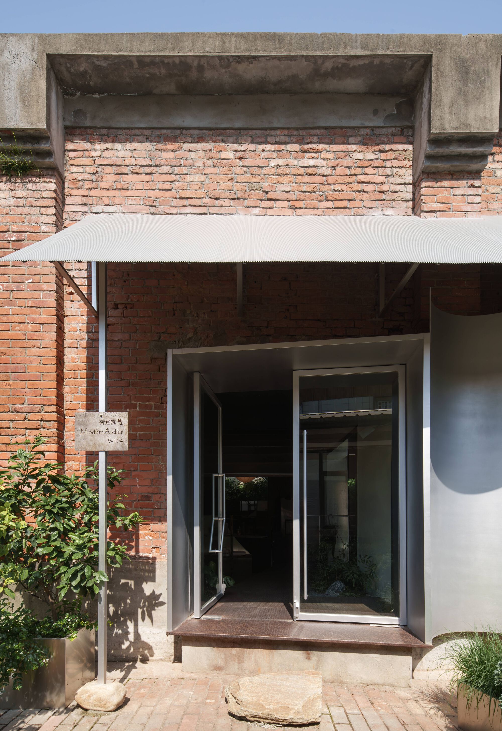Recessed entrance with exposed brick wall, concrete lintel and glazed pivot door under white canopy