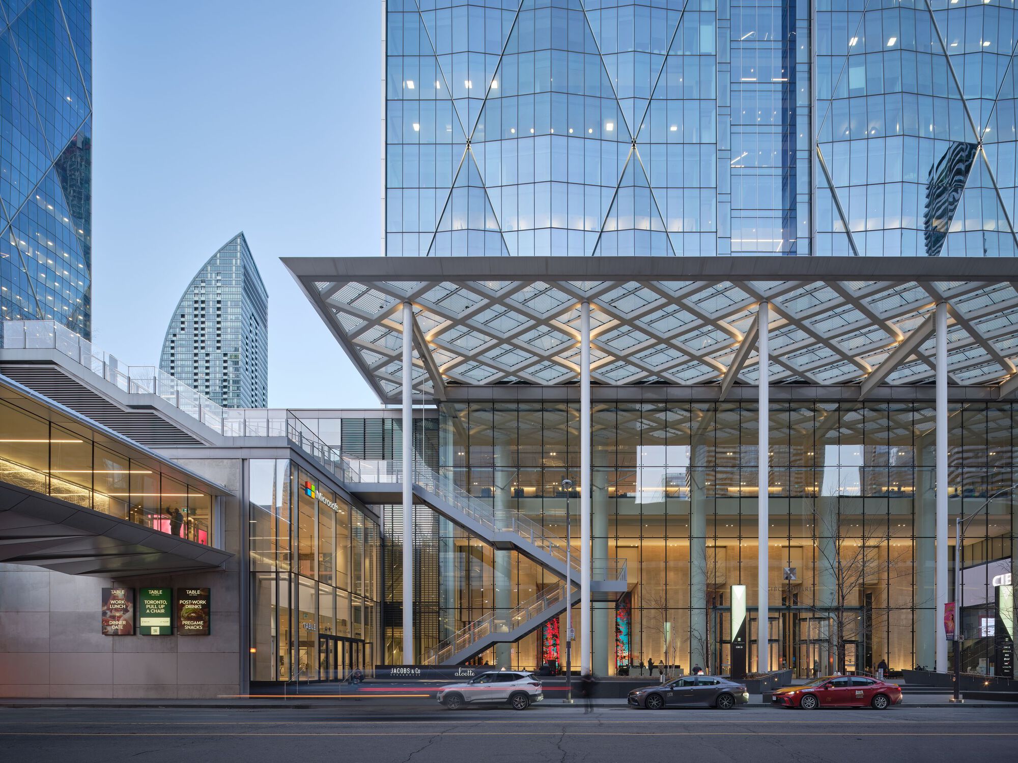 Street view of the glass pavilion entrance with diagonal lattice canopy and elevated pedestrian bridge